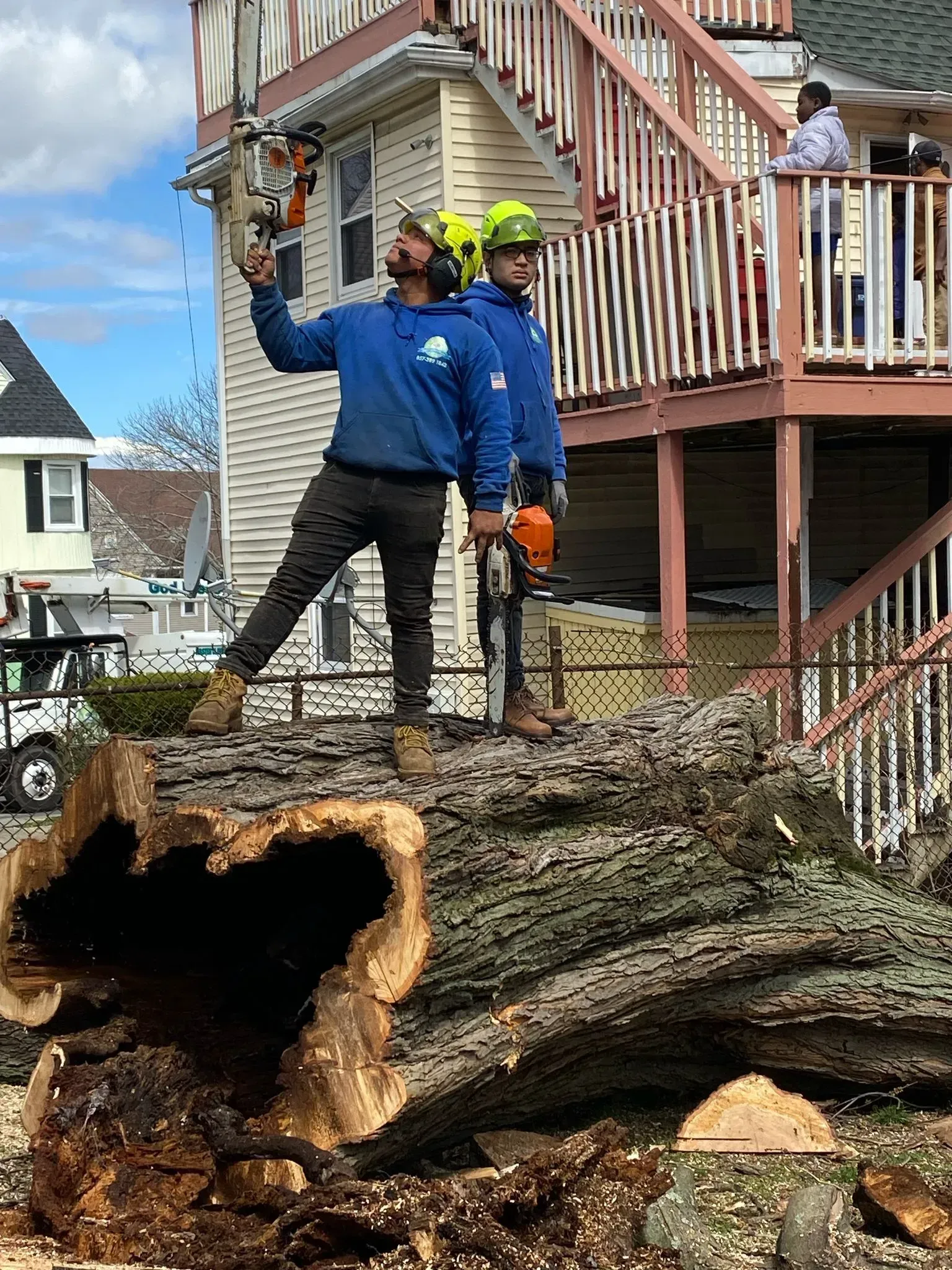 Workers in blue jackets and yellow helmets cut up a fallen tree beside a house and porch