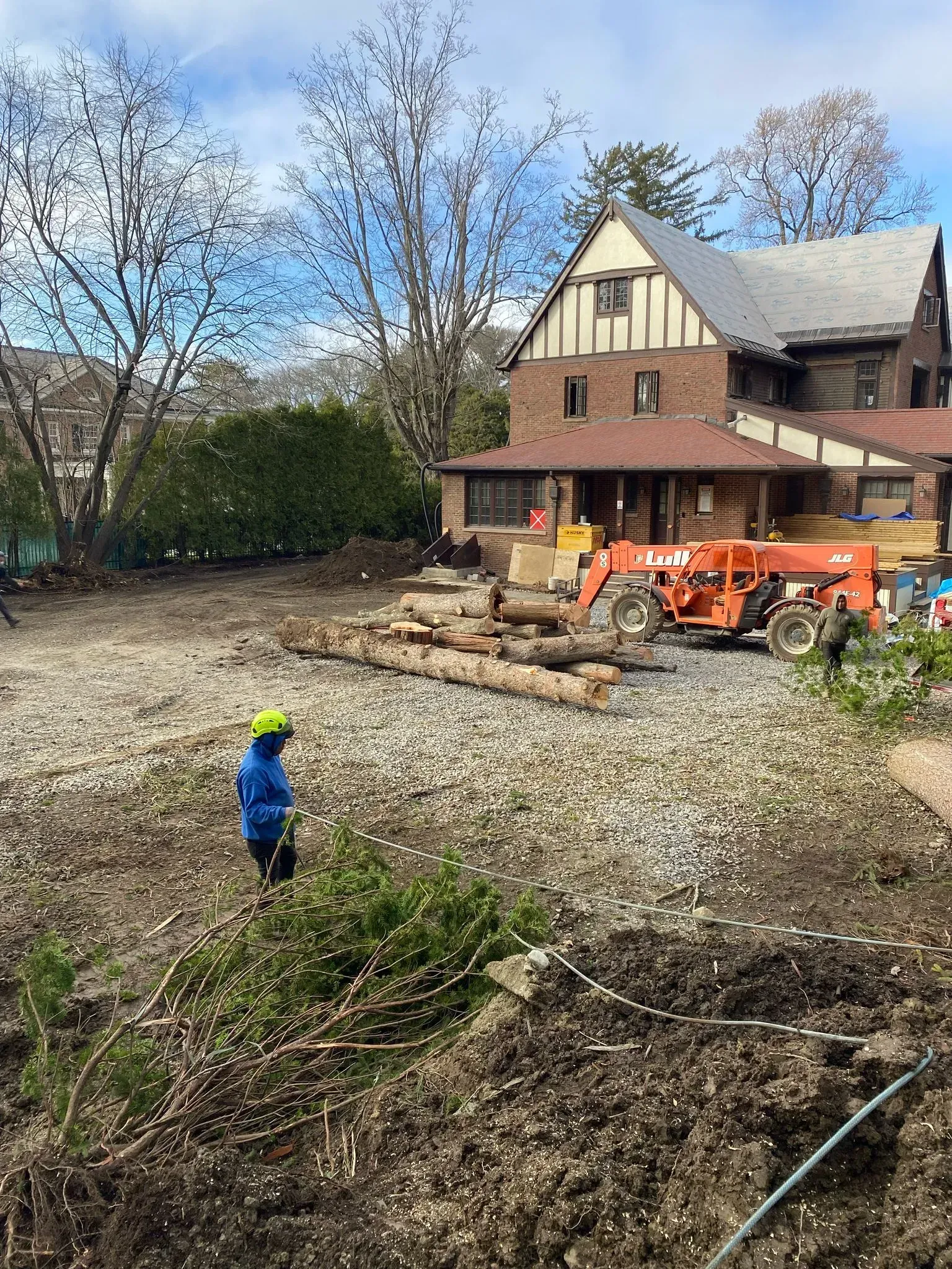Construction site with excavator, log piles, and a brick house under renovation, worker in blue in foreground