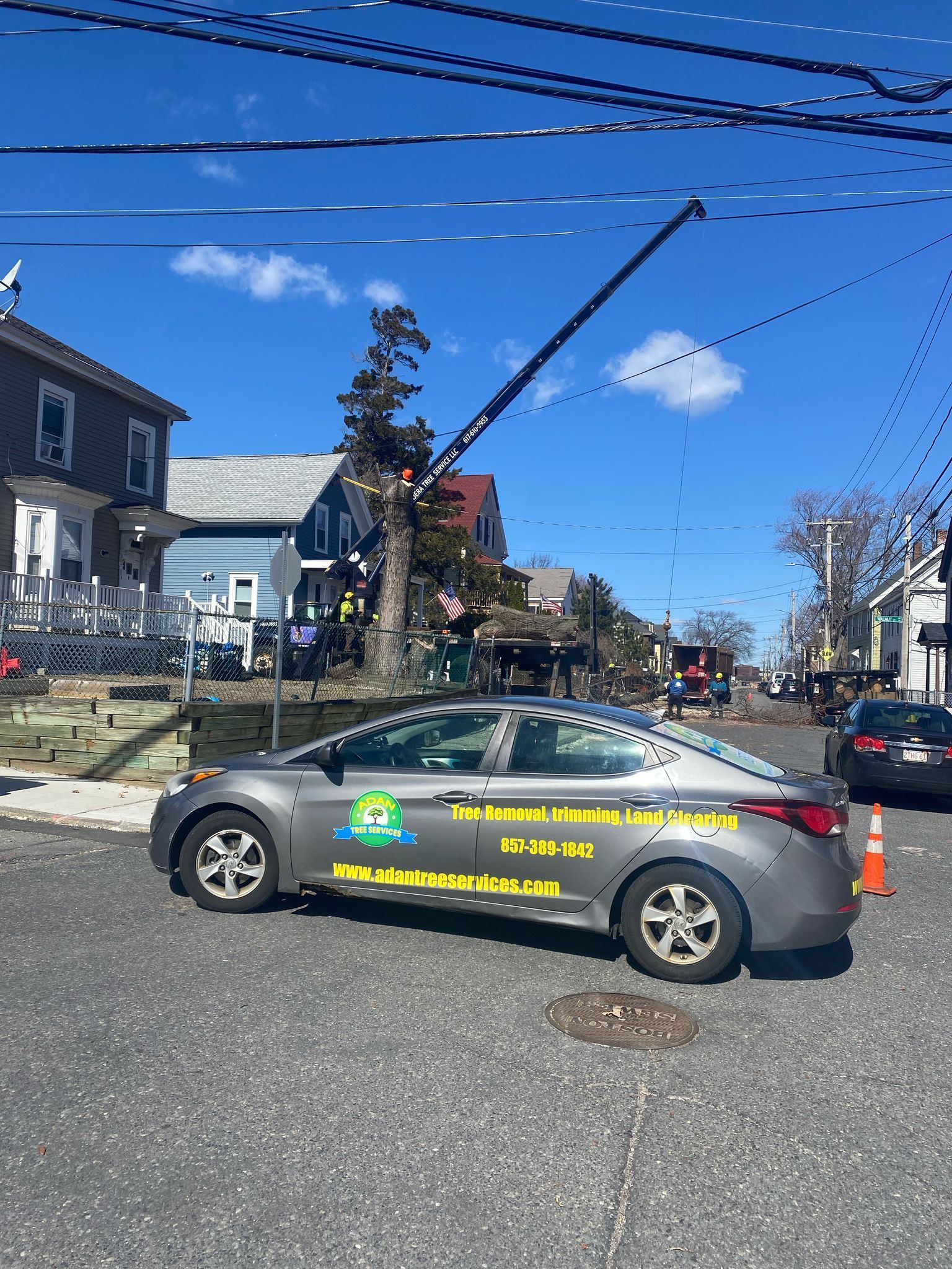 Gray utility car with yellow lettering parked on a street near a tree and houses, under a blue sky