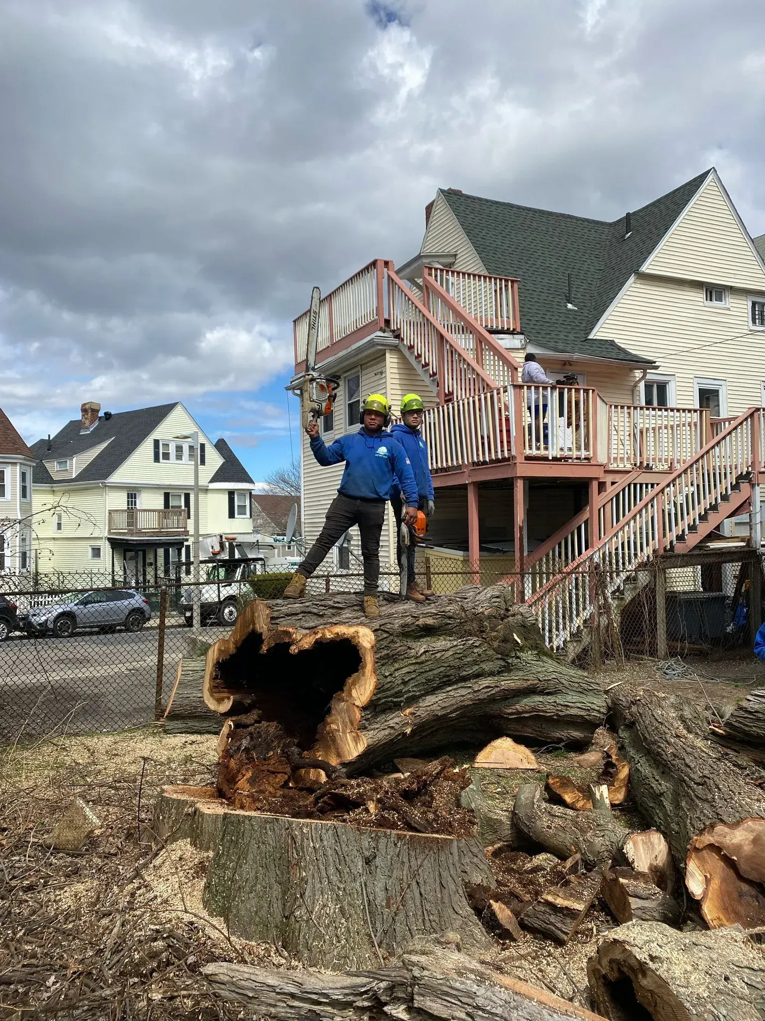 Two workers stand by a collapsed tree trunk and damaged deck beneath a house in a storm-damaged neighborhood.