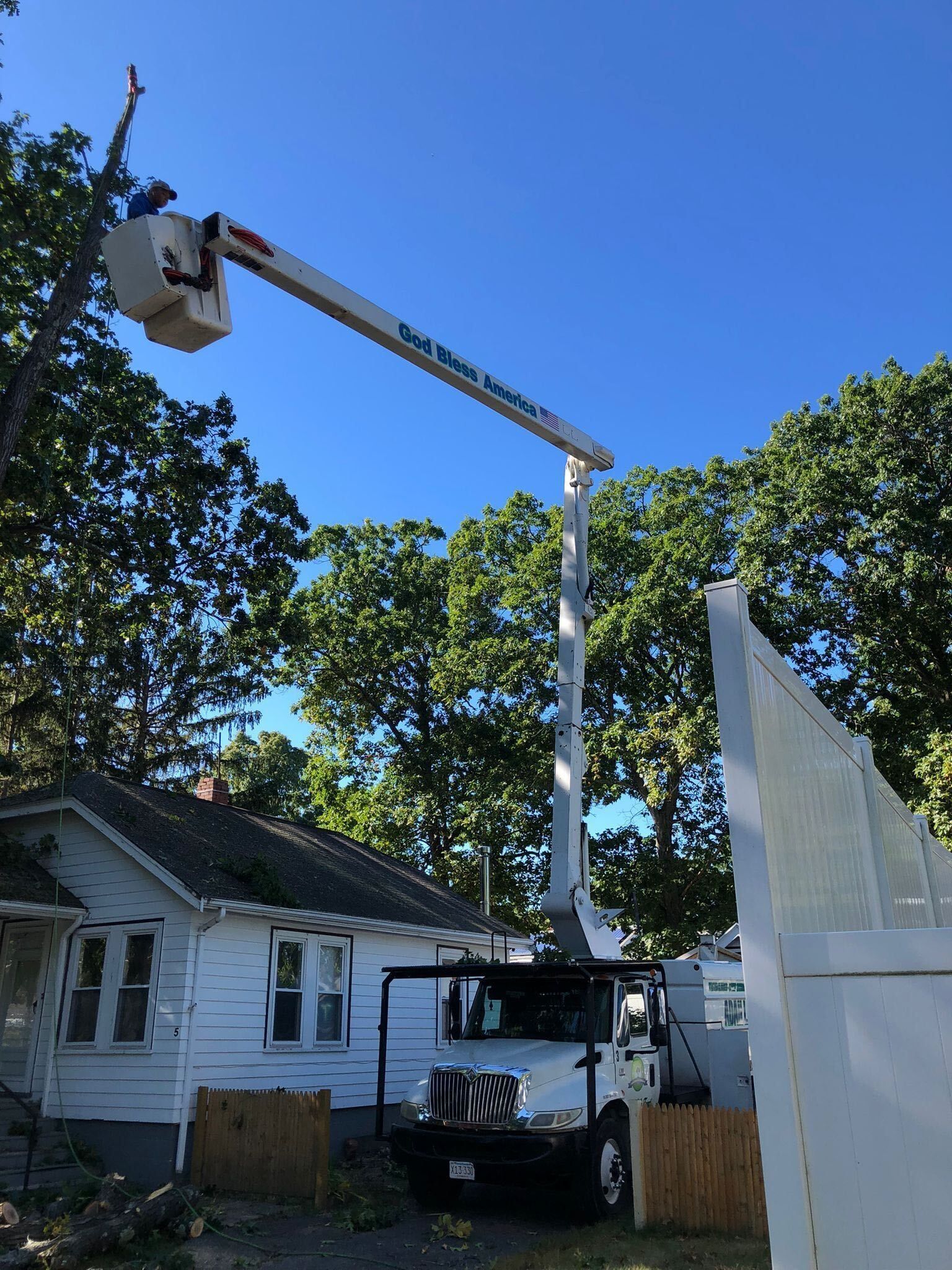Bucket truck raised beside a white house, with a worker basket near tree branches and a tall fence.