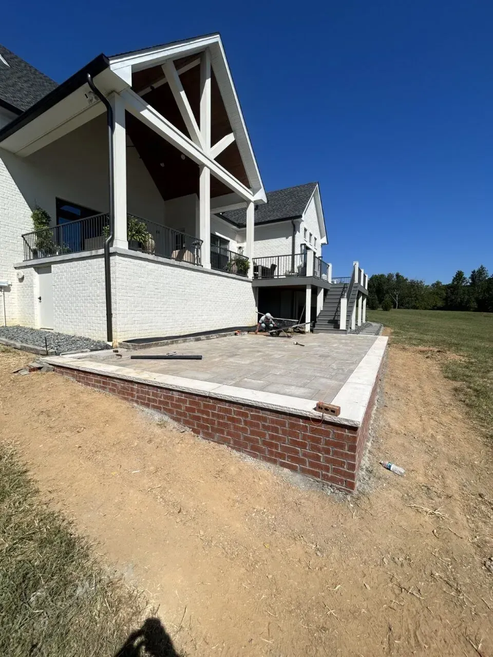 A brick patio being constructed next to a white house with a covered porch and deck on a sunny day.