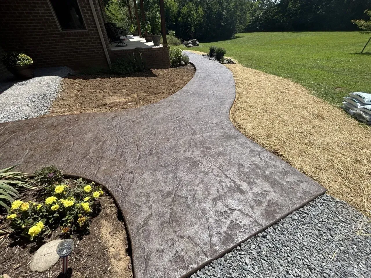 Brown stamped concrete pathway curves through a yard, bordered by gravel, mulch, and grass.