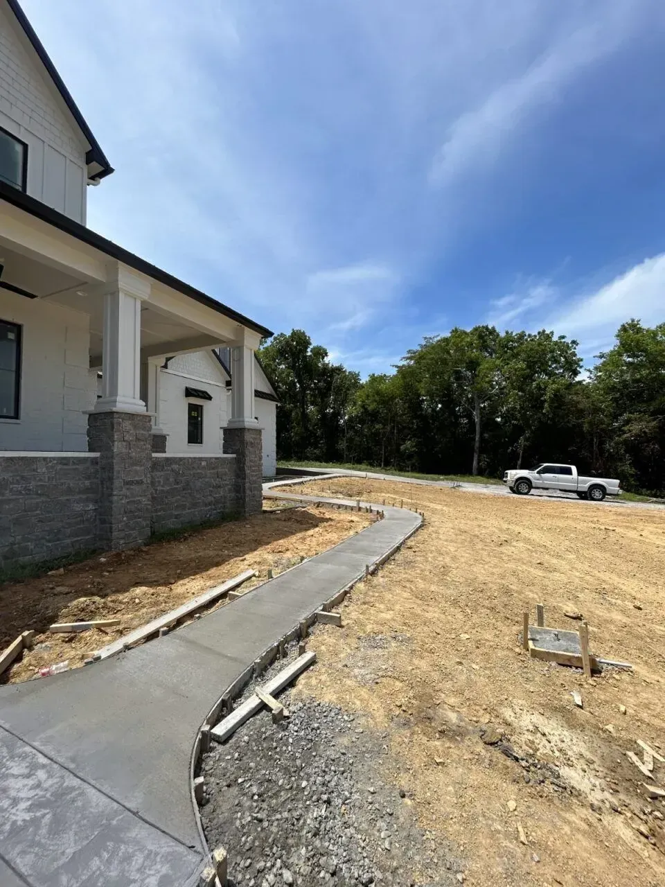 New concrete walkway leading to a house with white siding and gray stone accents; a truck is parked in the distance.