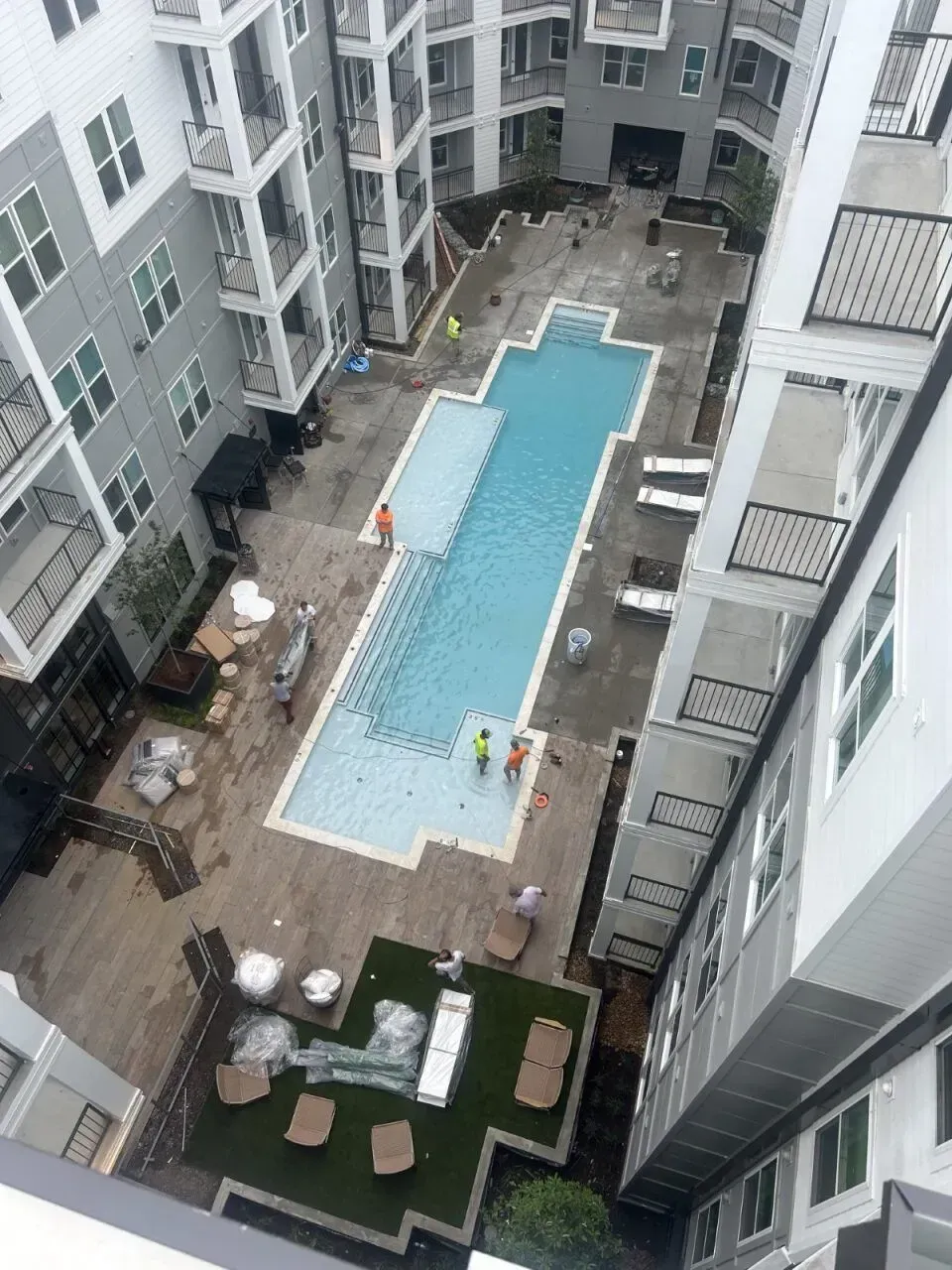 Aerial view of a rectangular swimming pool surrounded by a multi-story apartment complex. Several people are present, possibly working.