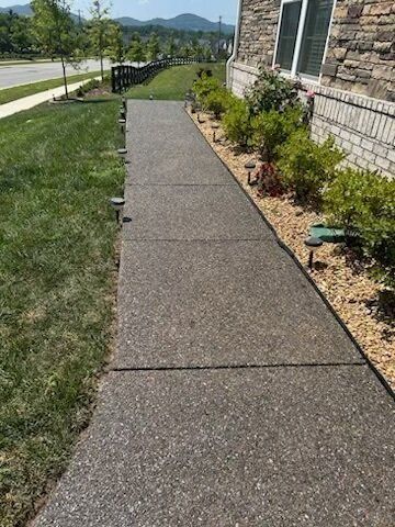 Concrete walkway flanked by grass and landscaping leading to a stone house.