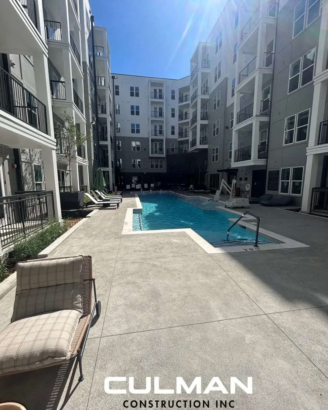 Pool area in apartment complex, concrete patio, lounge chairs, blue water, multi-story buildings.