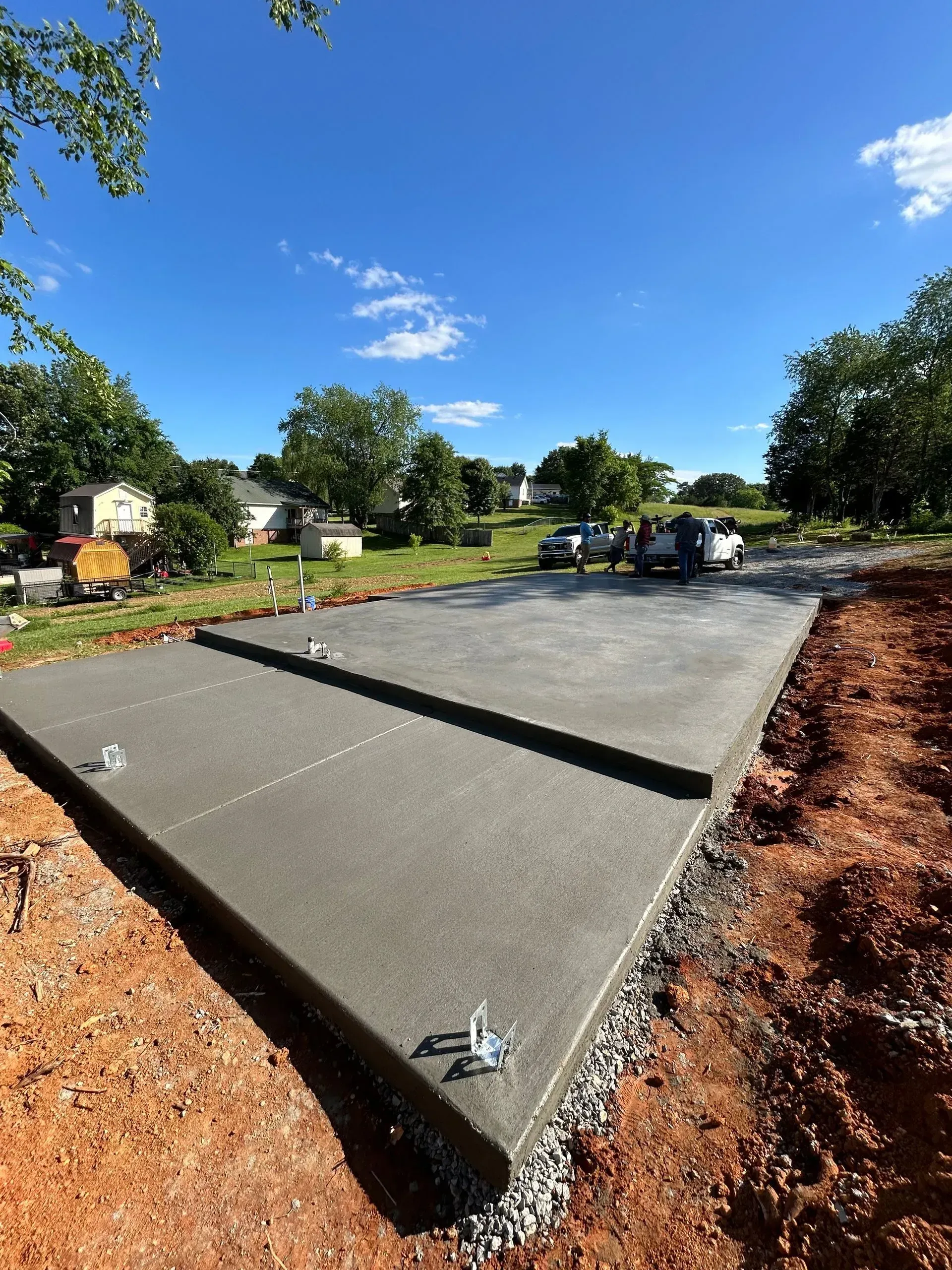Freshly poured concrete slab with embedded anchors, surrounded by dirt and gravel. Blue sky, trees, and houses in the background.