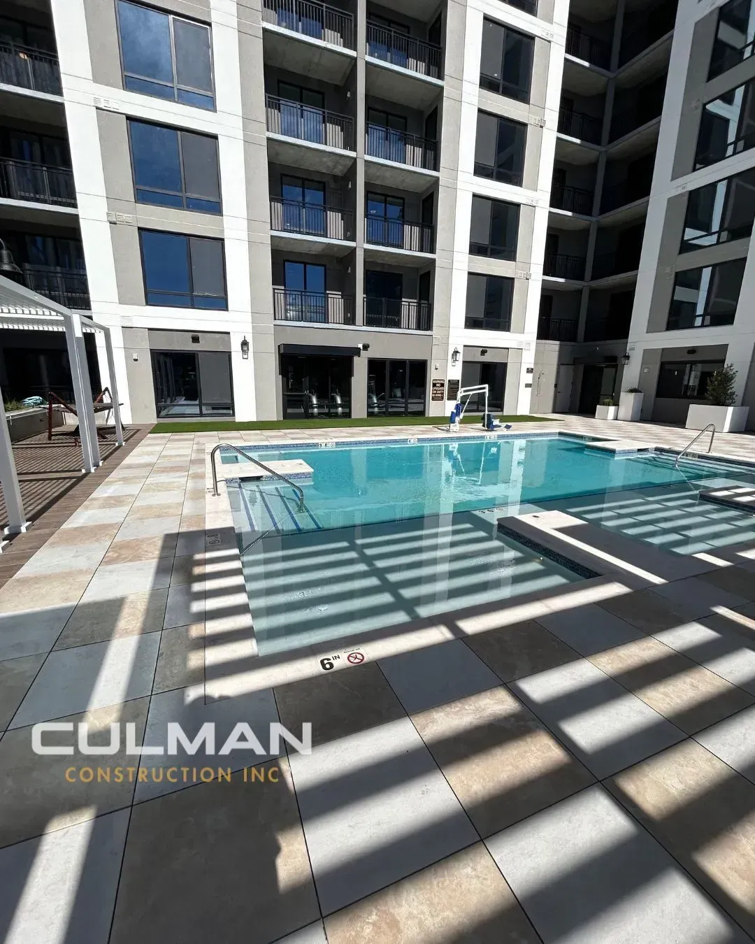 Pool and patio area in front of a modern apartment building. Beige tile, clear water, and sunny conditions.