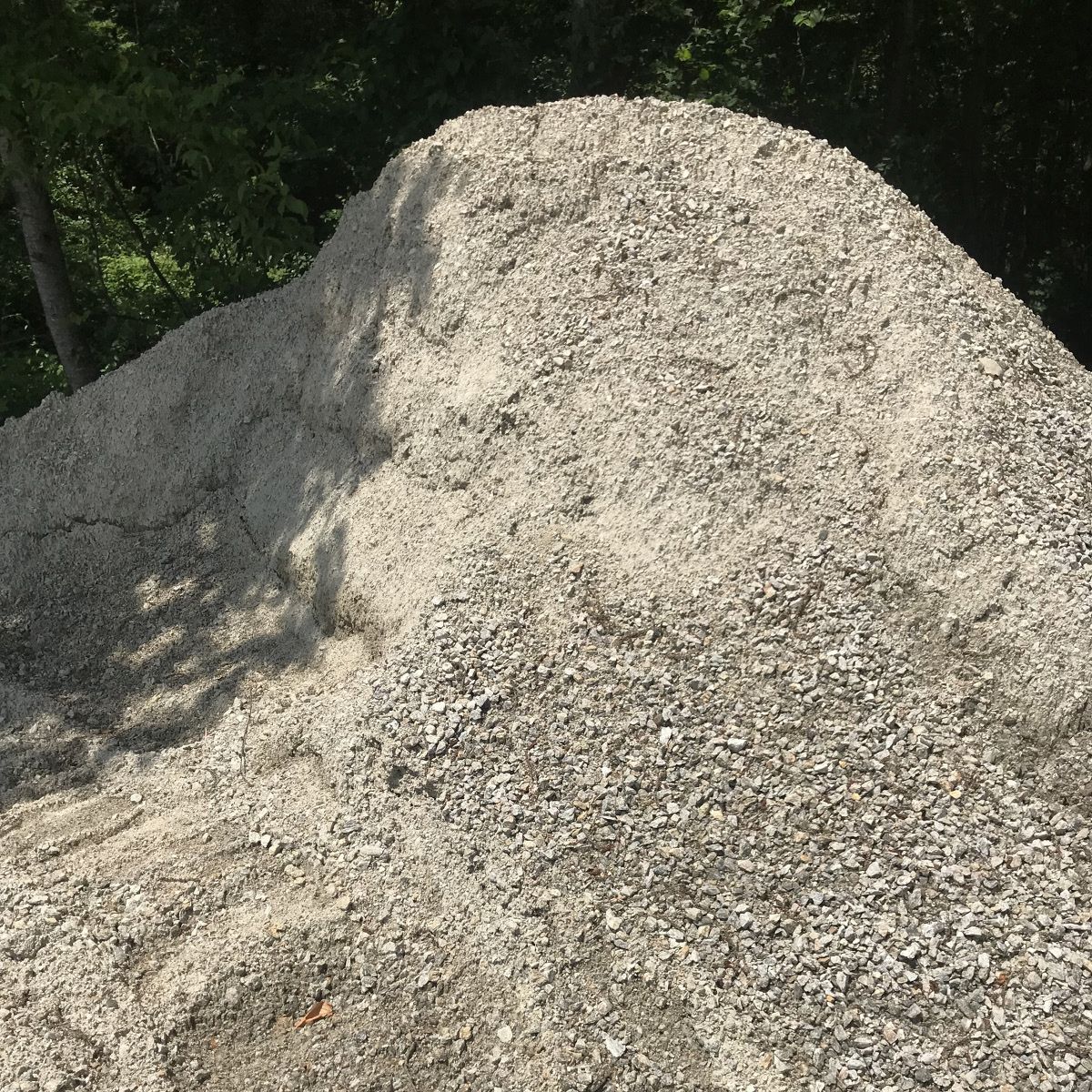 A pile of gravel is sitting in the shade of trees.