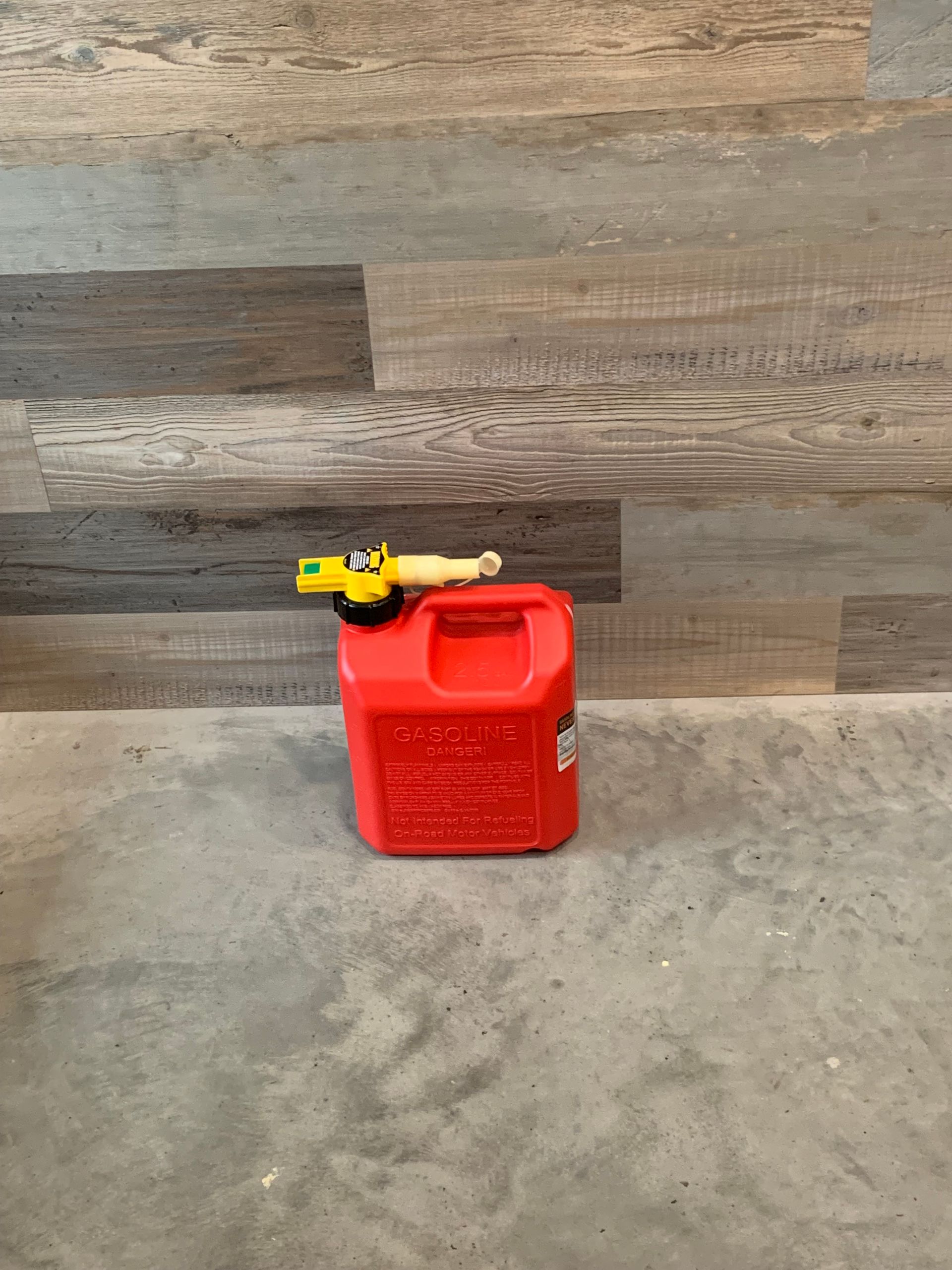 A red gas can is sitting on a concrete floor in front of a wooden wall.