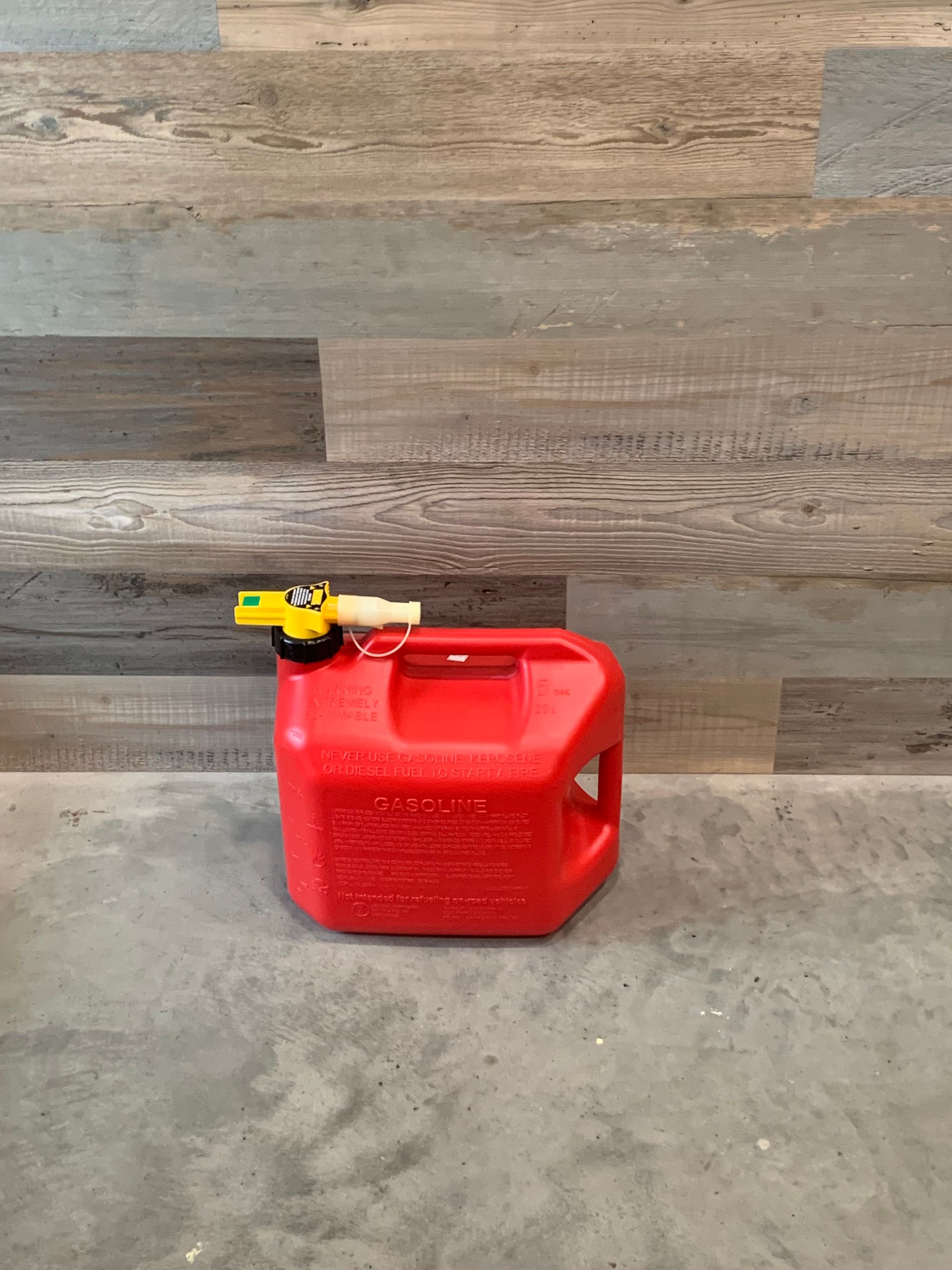 A red gas can is sitting on a concrete floor in front of a wooden wall.