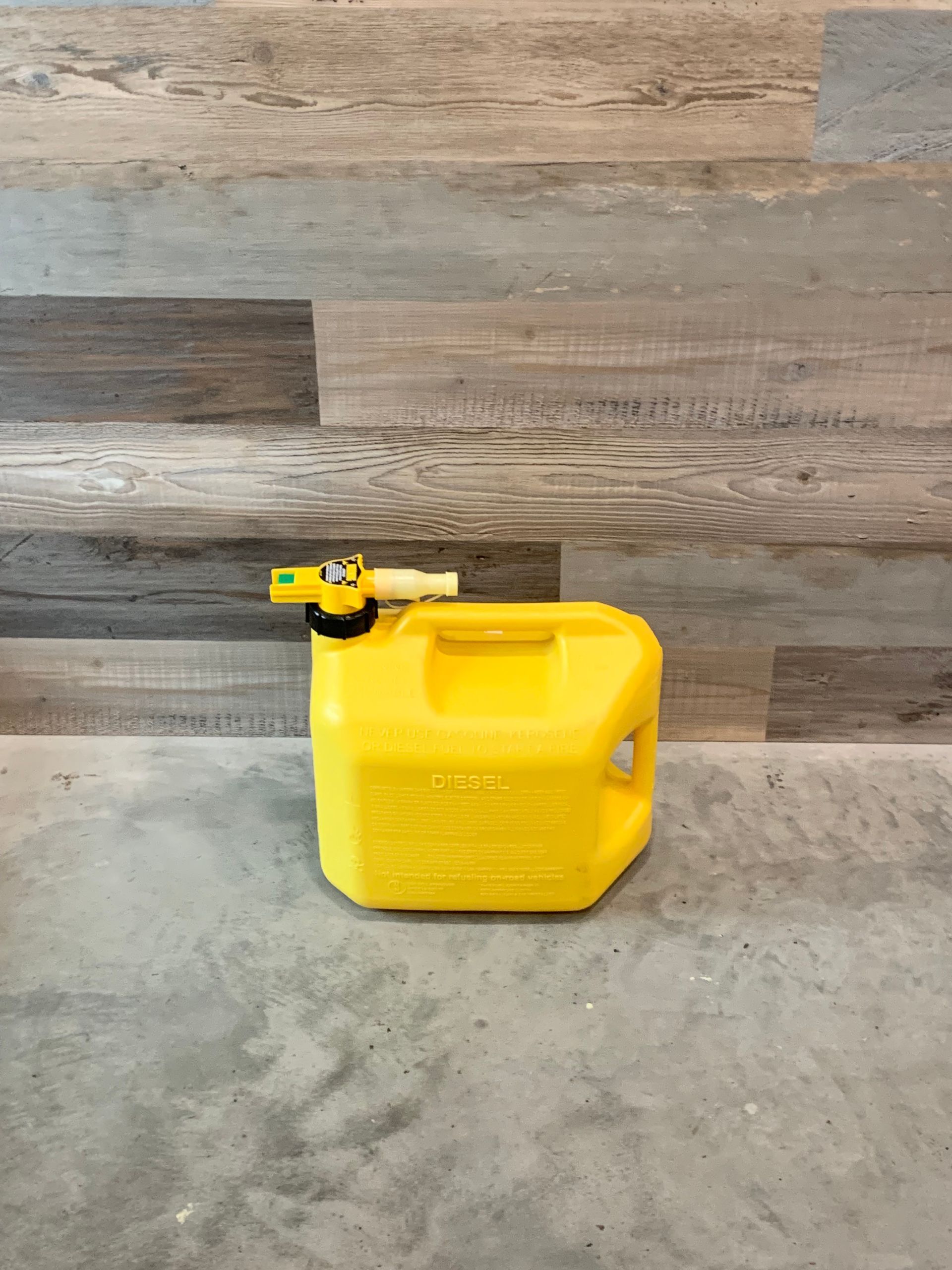 A yellow gas can is sitting on a concrete floor in front of a wooden wall.