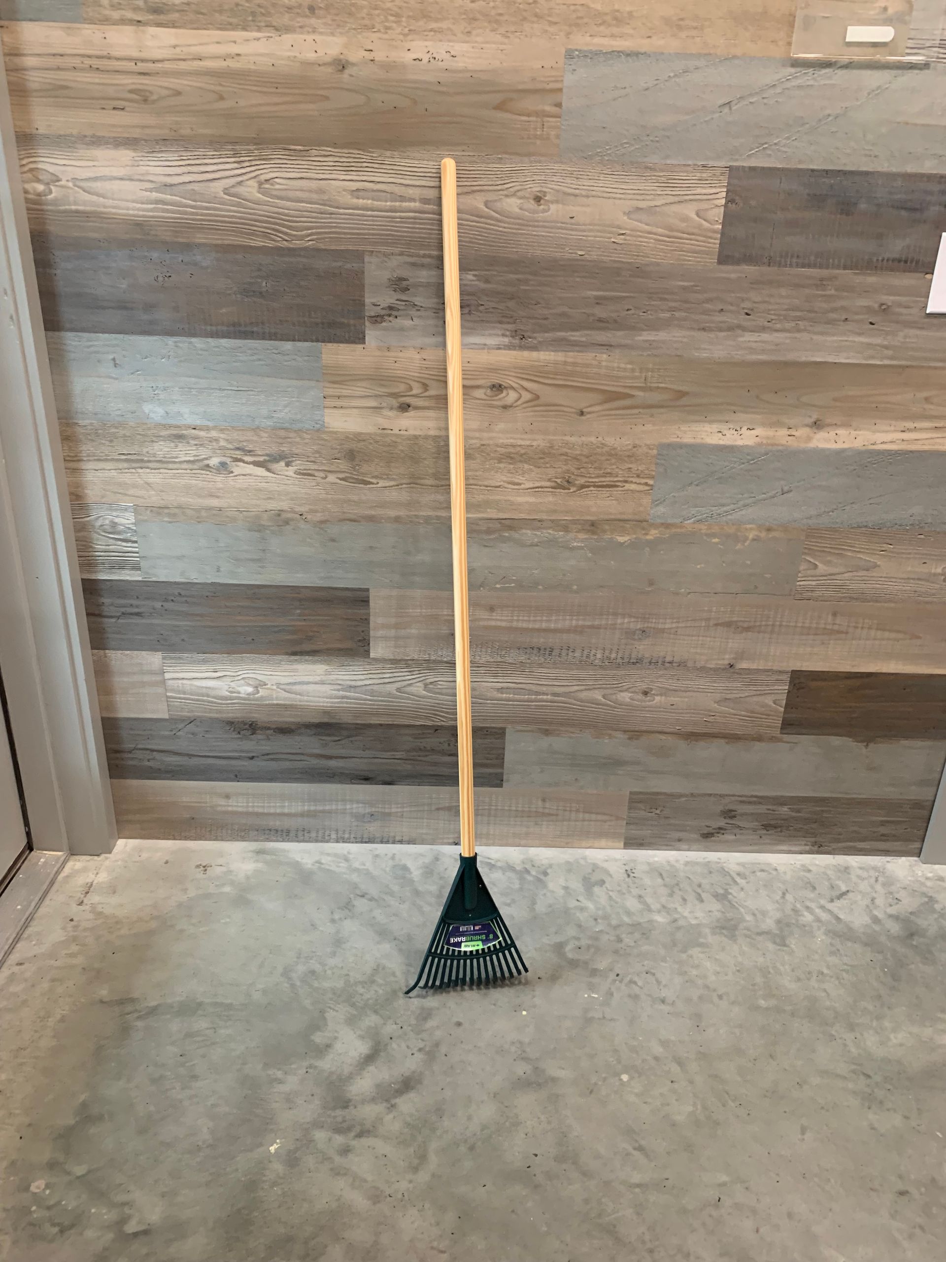 A green rake with a wooden handle is sitting on a concrete floor in front of a wooden wall.