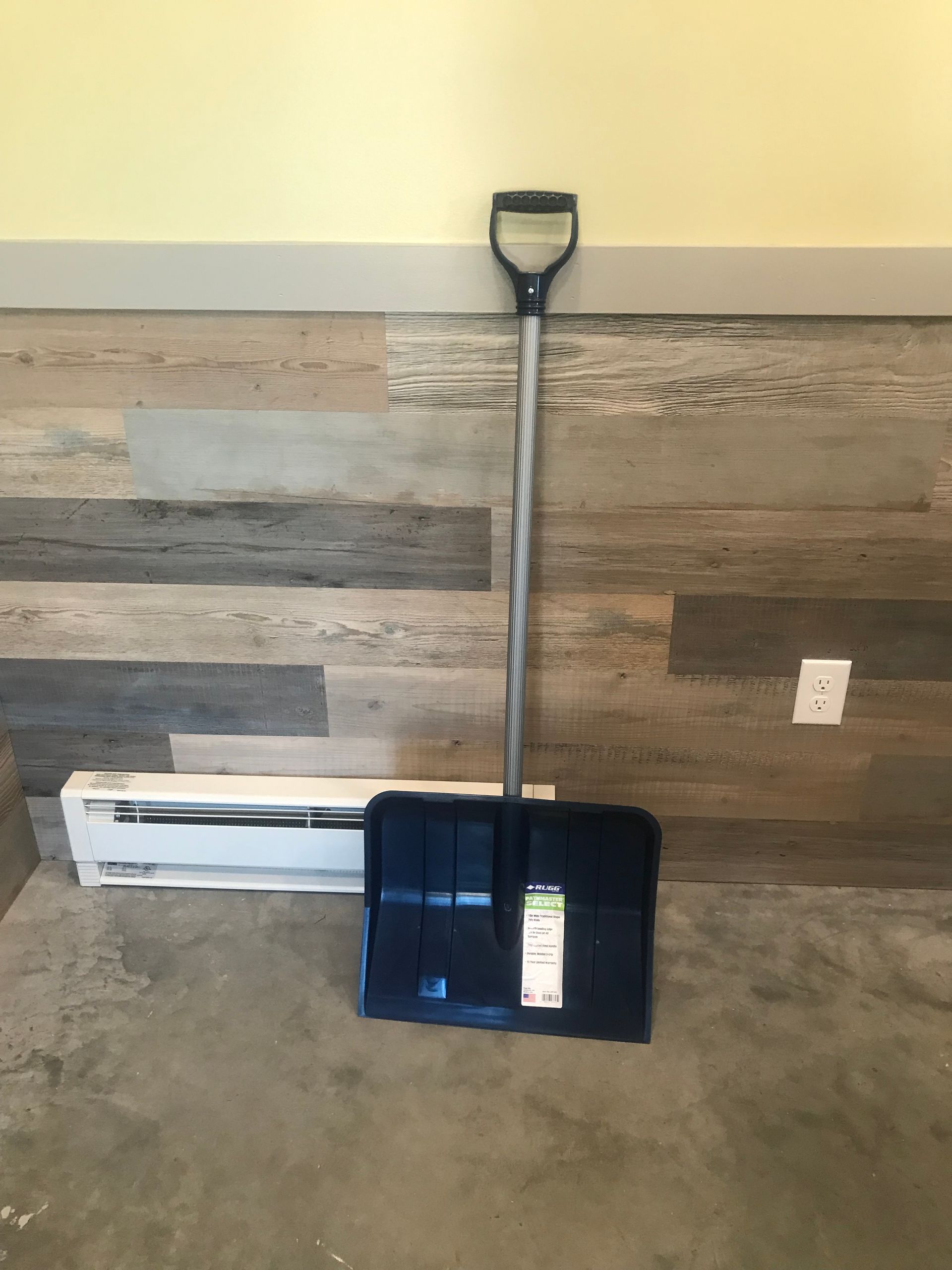 A snow shovel is sitting on the floor in front of a wooden wall.
