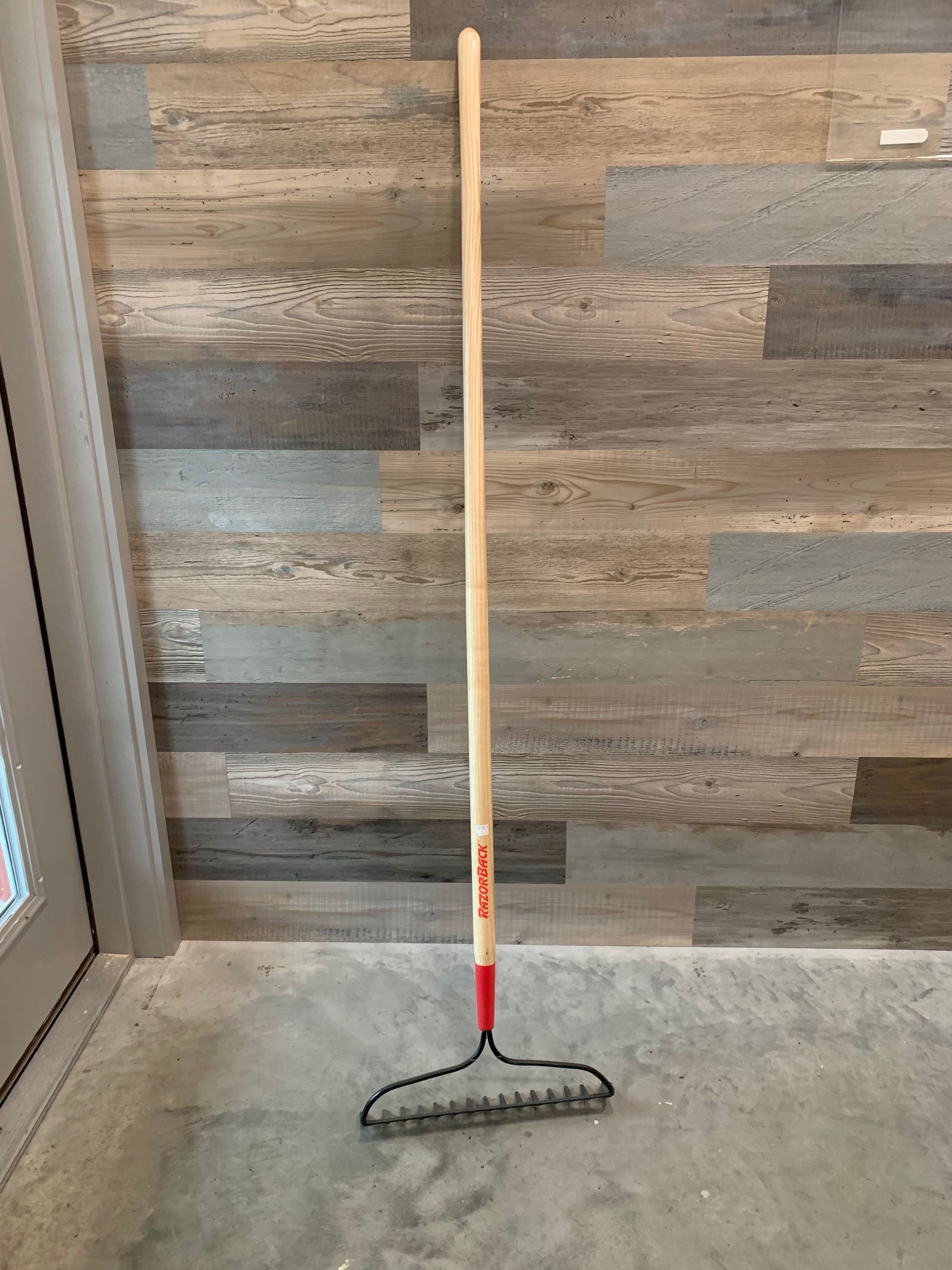 A rake with a wooden handle is sitting on the floor in front of a wooden wall.