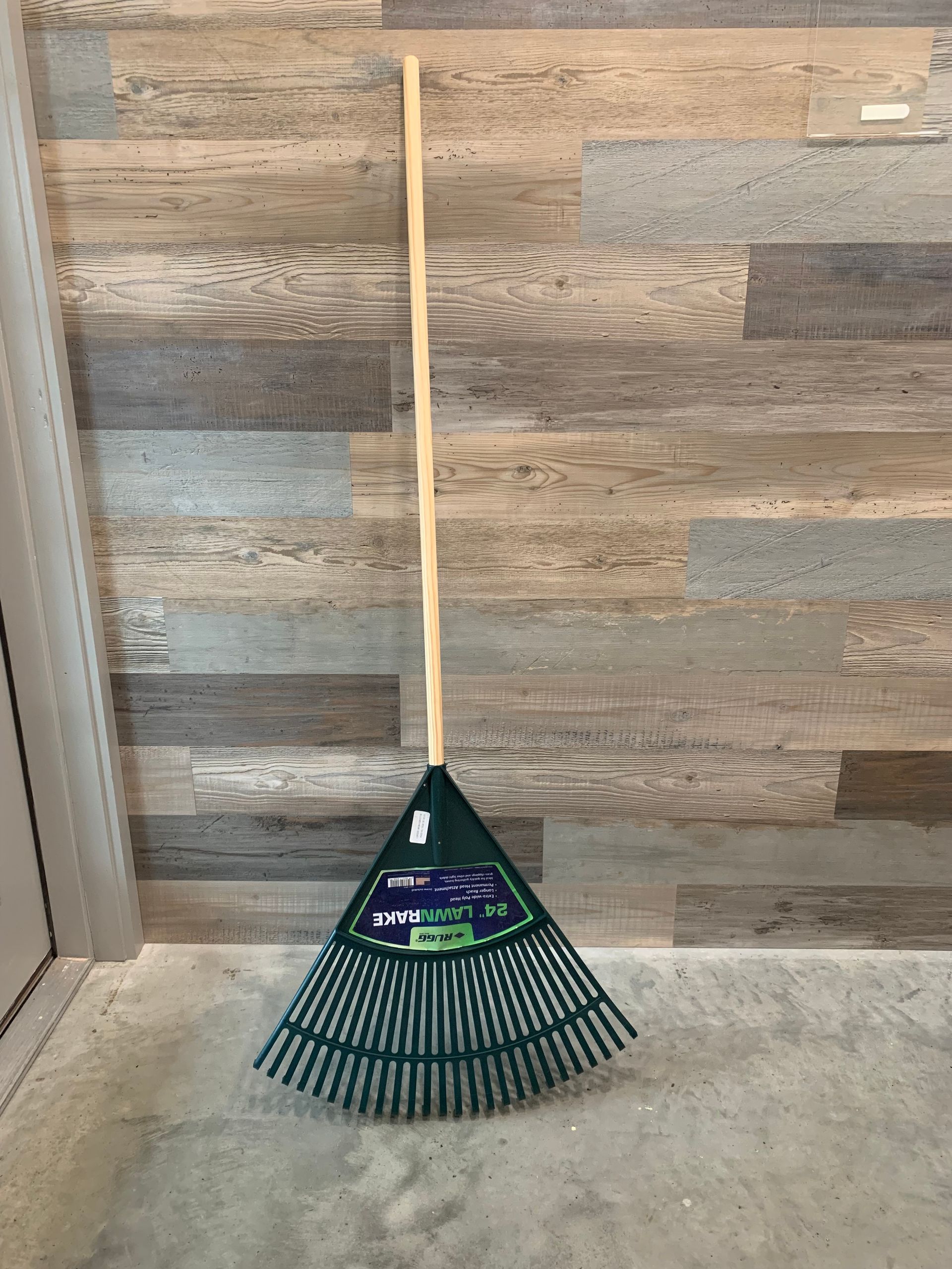 A green rake with a wooden handle is sitting on the floor in front of a wooden wall.