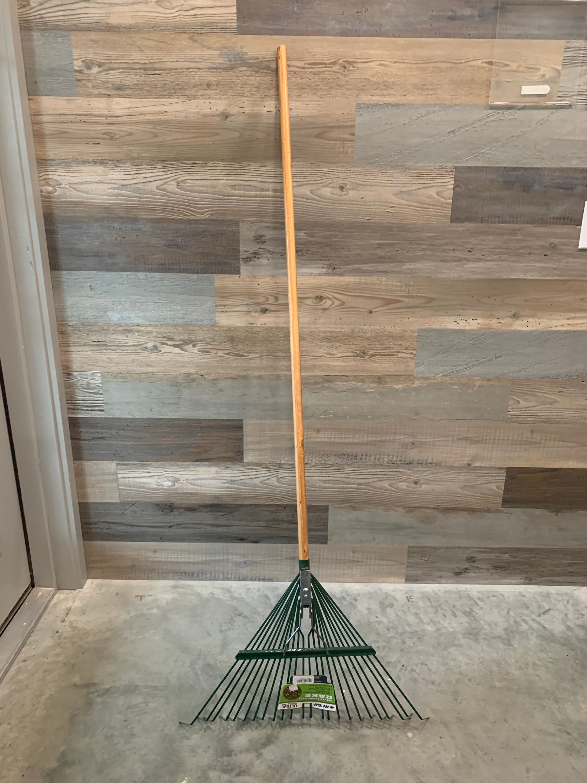 A green rake with a wooden handle is sitting on the floor in front of a wooden wall.