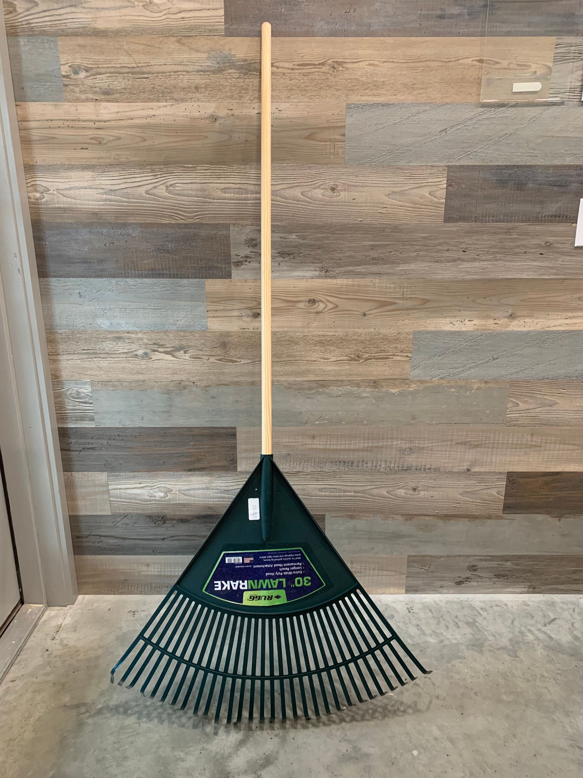 A green rake with a wooden handle is sitting on the floor in front of a wooden wall.