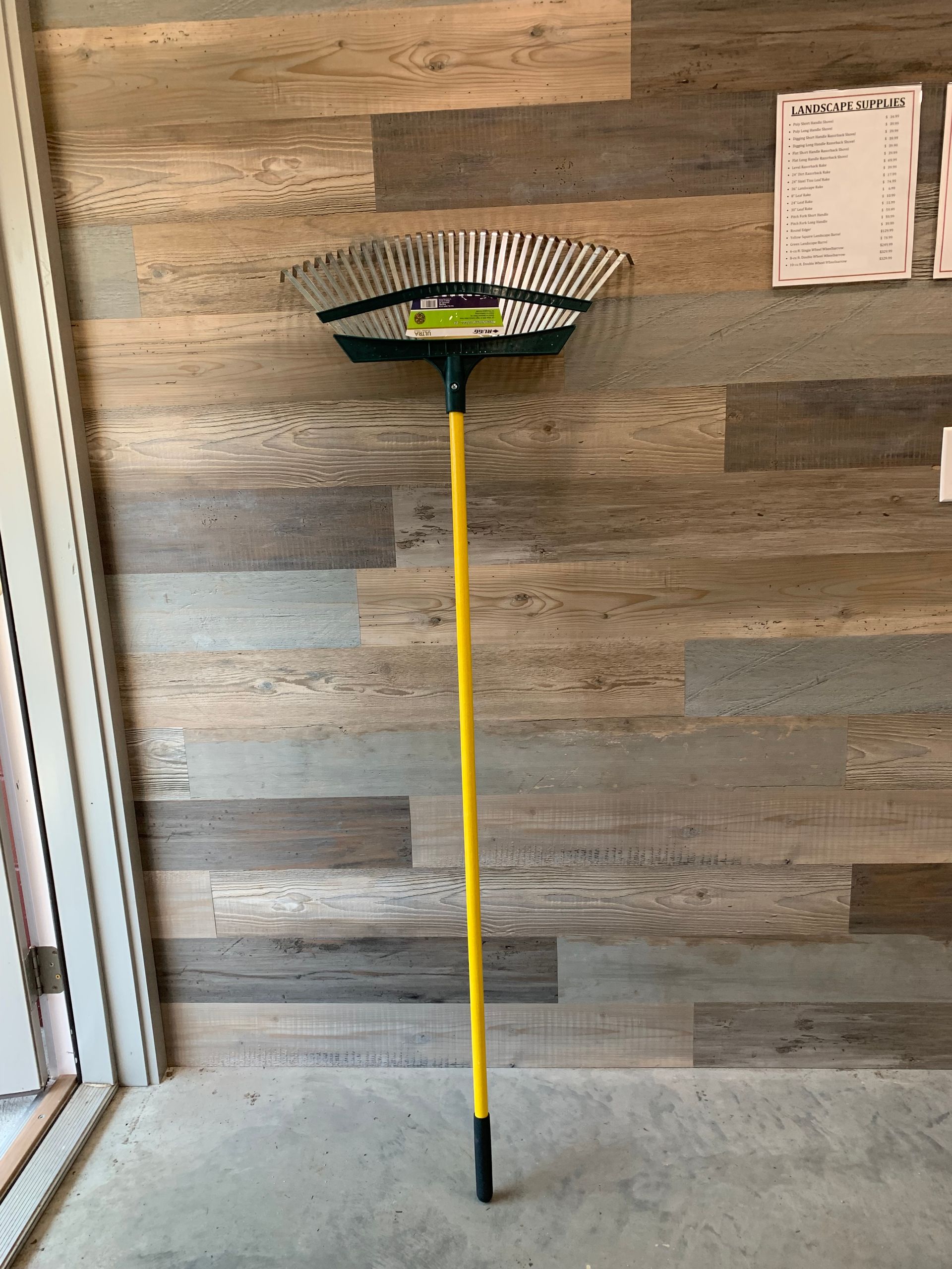 A rake with a yellow handle is sitting in front of a wooden wall.