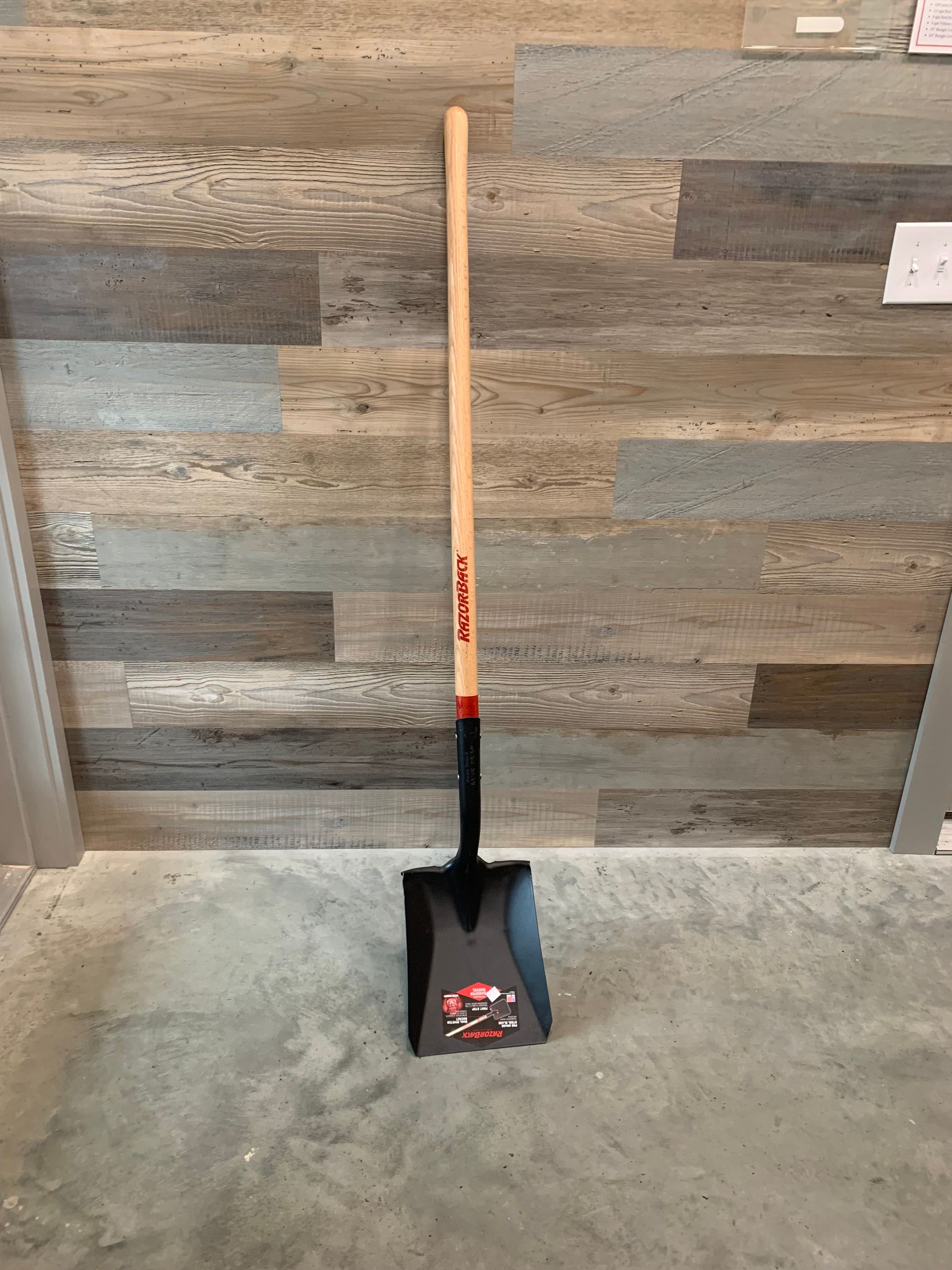 A shovel with a wooden handle is sitting on a concrete floor in front of a wooden wall.
