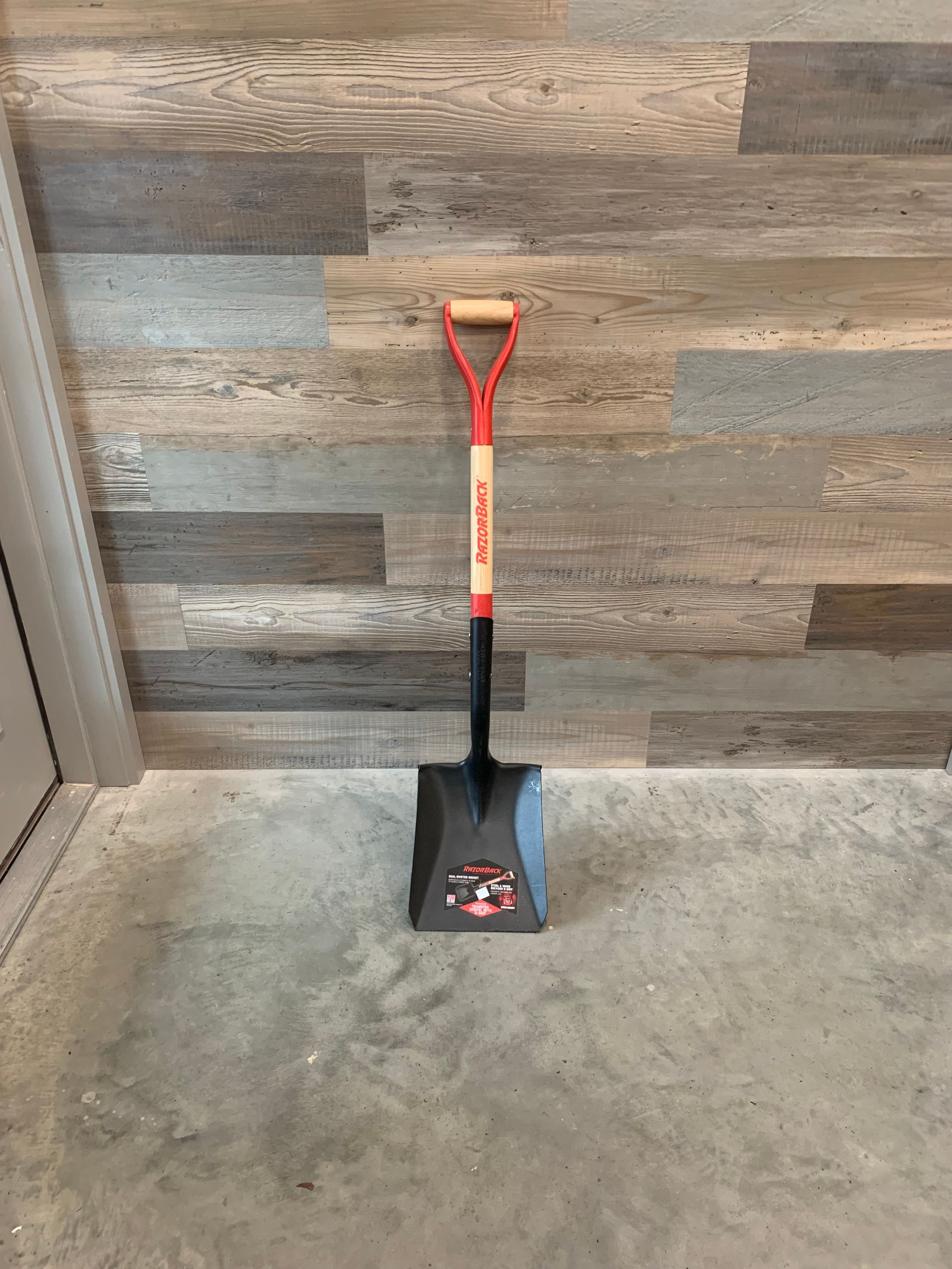 A shovel is sitting on a concrete floor in front of a wooden wall.