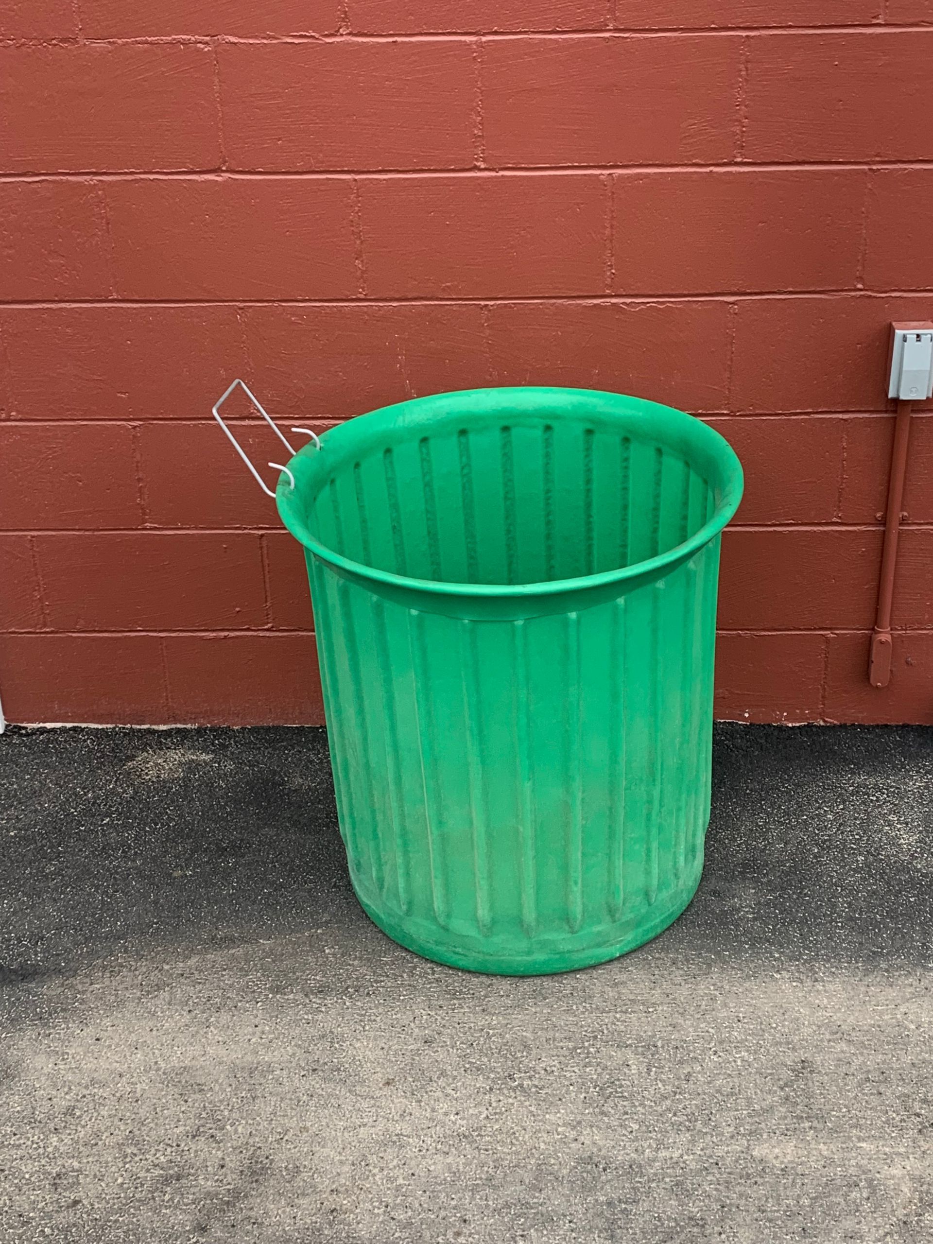 A green trash can is sitting in front of a red brick wall
