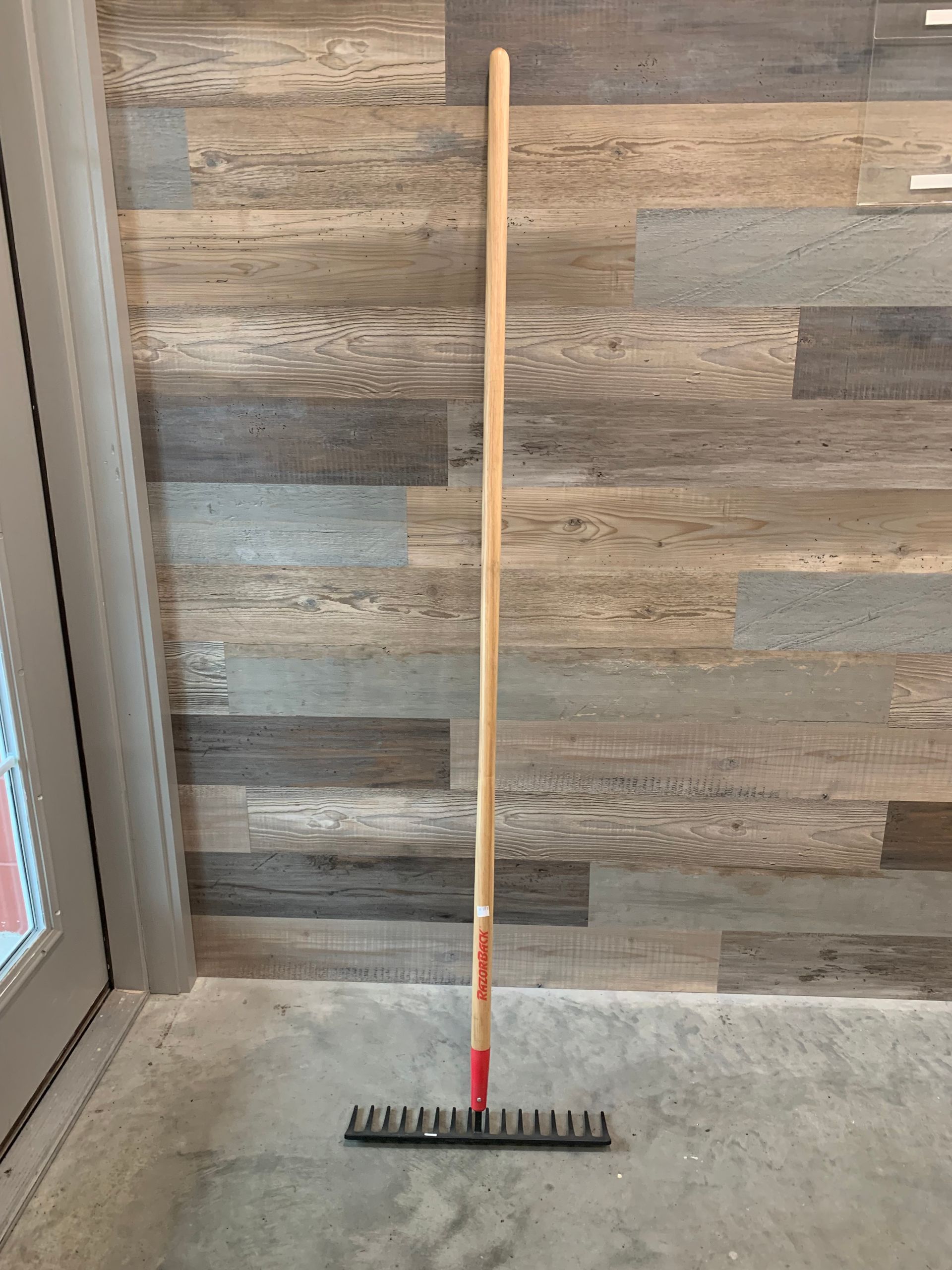 A rake with a wooden handle is sitting on the floor in front of a wooden wall.
