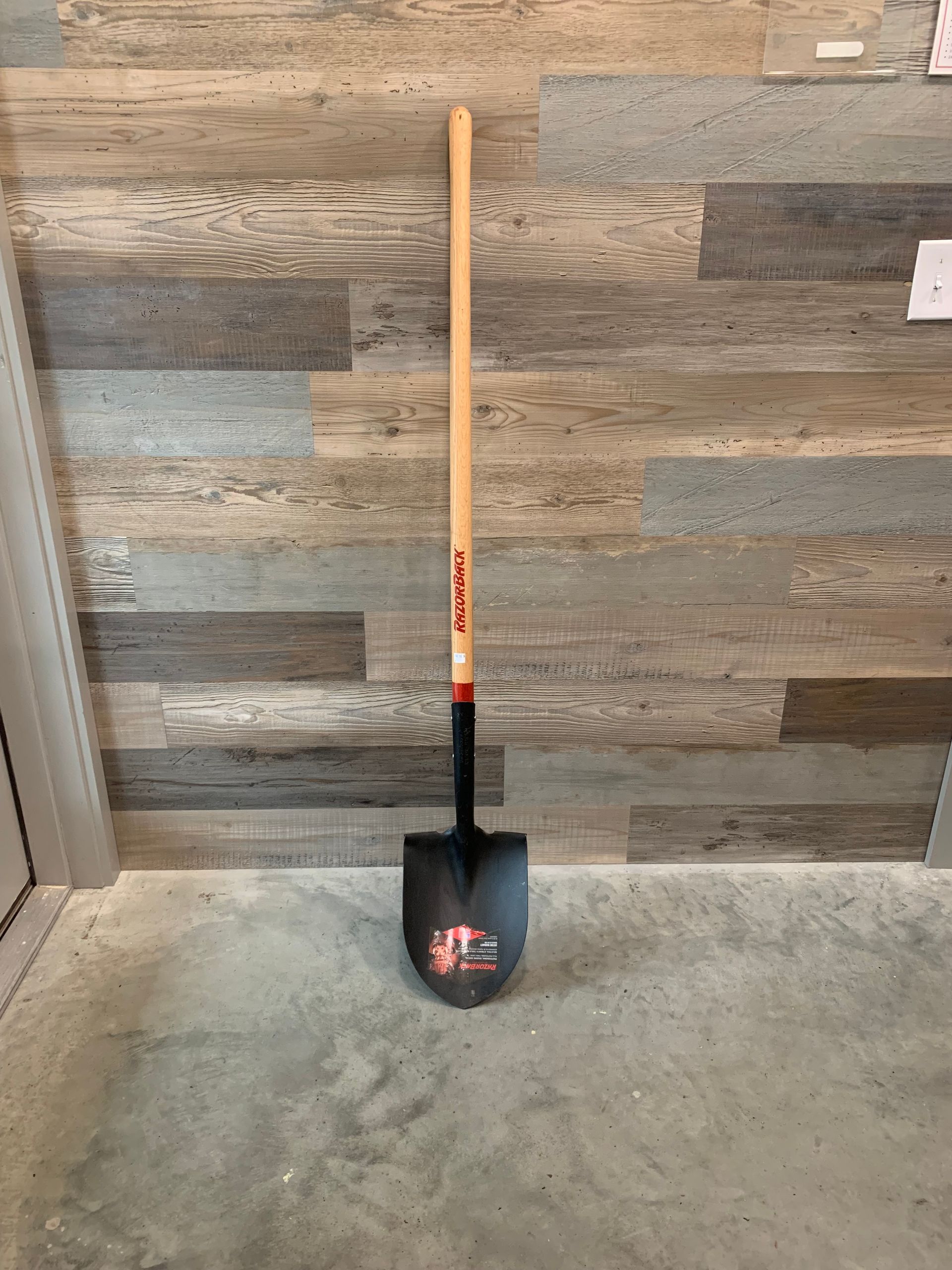 A shovel with a wooden handle is sitting on the floor in front of a wooden wall.