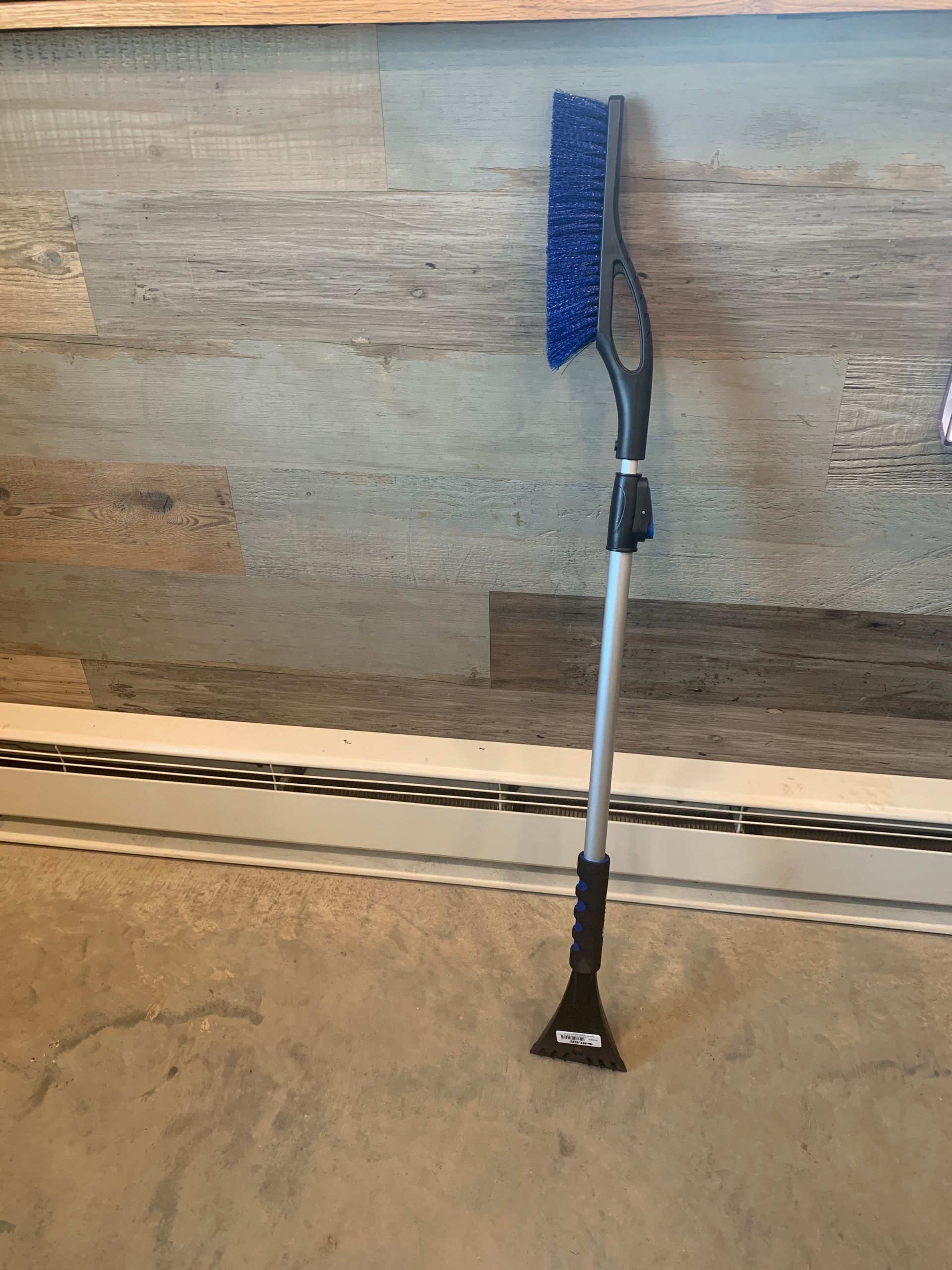 A snow broom is sitting on the floor next to a wooden wall.