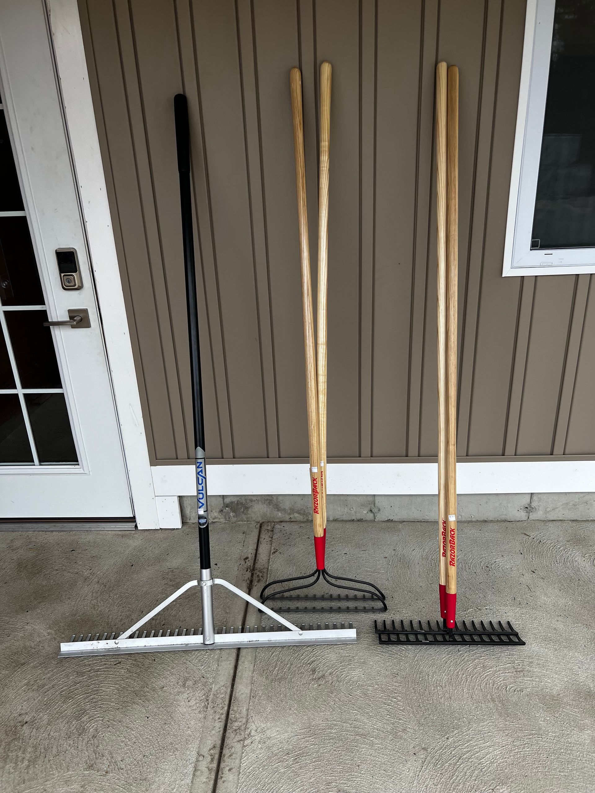 Three rakes are sitting on the sidewalk in front of a house.