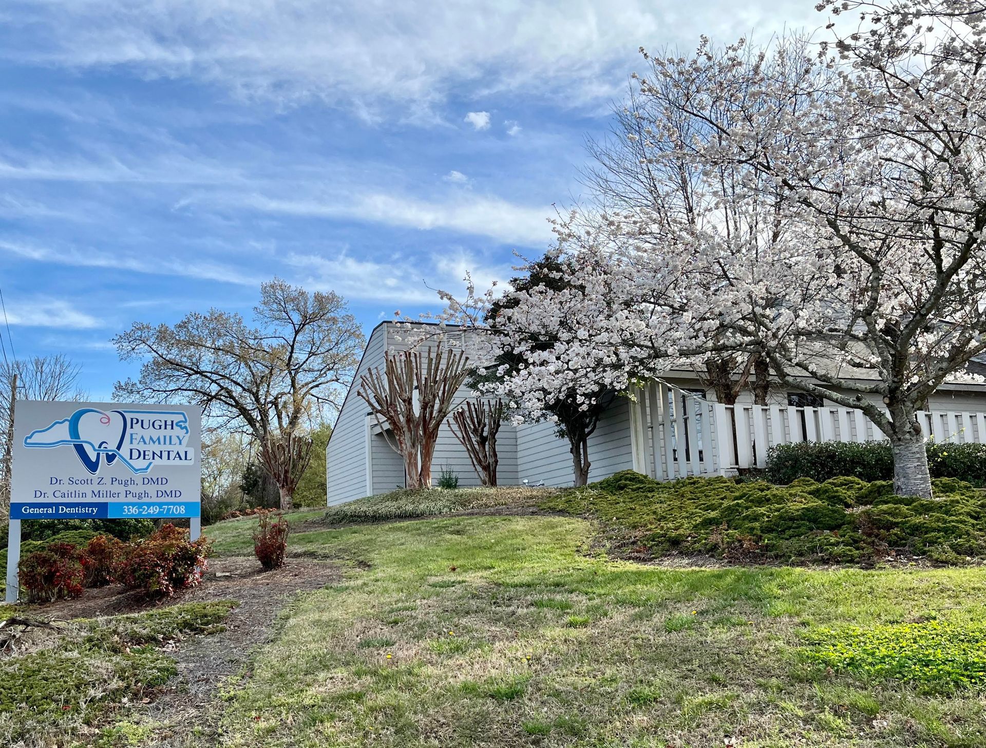 White building with blooming trees and a blue sign in a grassy front yard under a partly cloudy sky