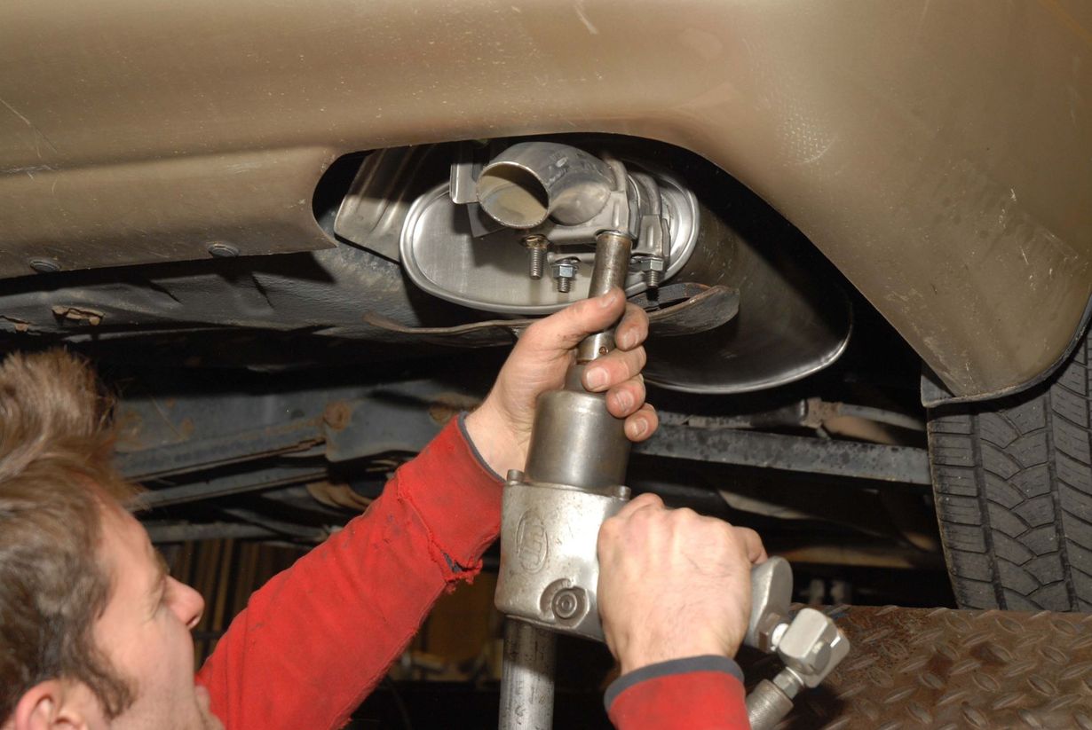 Mechanic using power tool on a car's exhaust system, viewed from below.