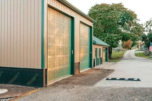 Two large tan garage doors with green trim, set in a light beige building. A gravel driveway leads away.