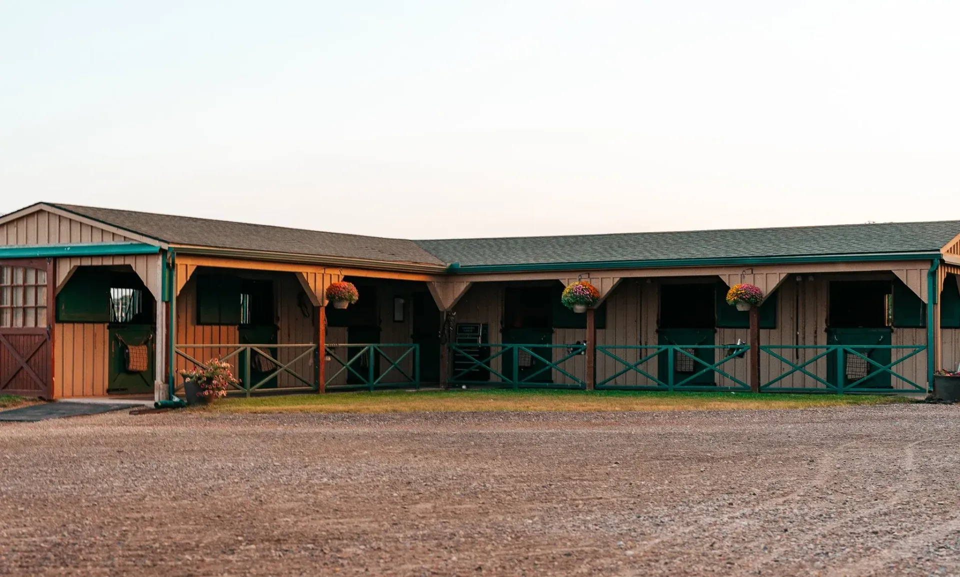 Horse stable with open stalls, beige and green trim, hanging flower baskets, gravel ground.