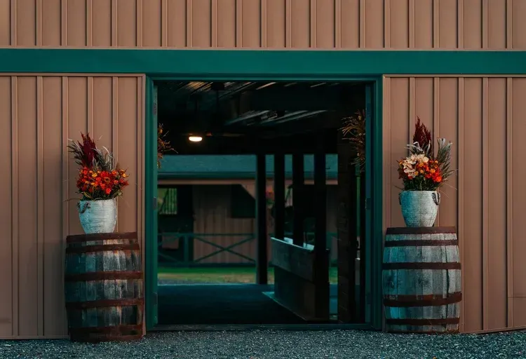 Exterior of a barn with open doorway. Flowers sit atop barrels on either side.