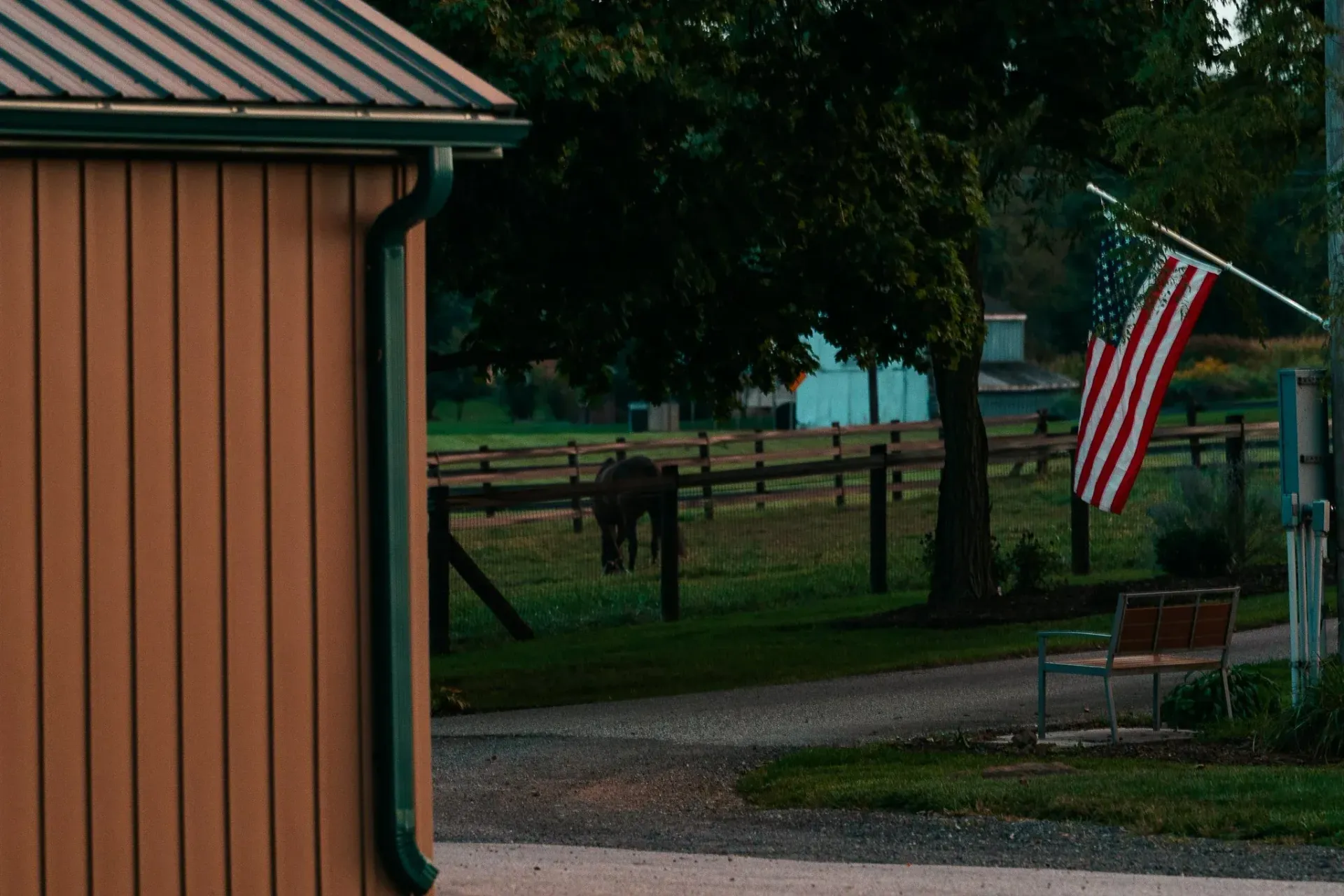 Brown building, American flag, horse grazing in a fenced field, gravel driveway, overcast.