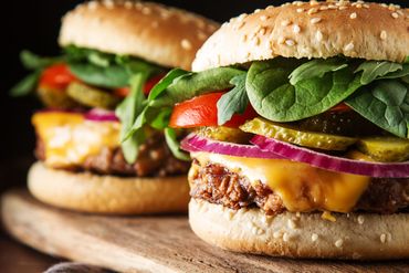 Two cheeseburgers on a wooden board. Sesame seed buns, layered with burger, cheese, pickles, red onion, tomato, and greens.