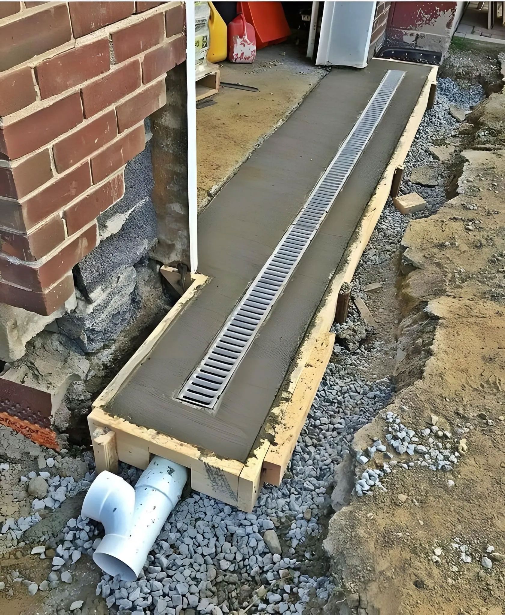 Trench drain with metal grate and concrete, connected to a PVC pipe, next to brick and gravel.