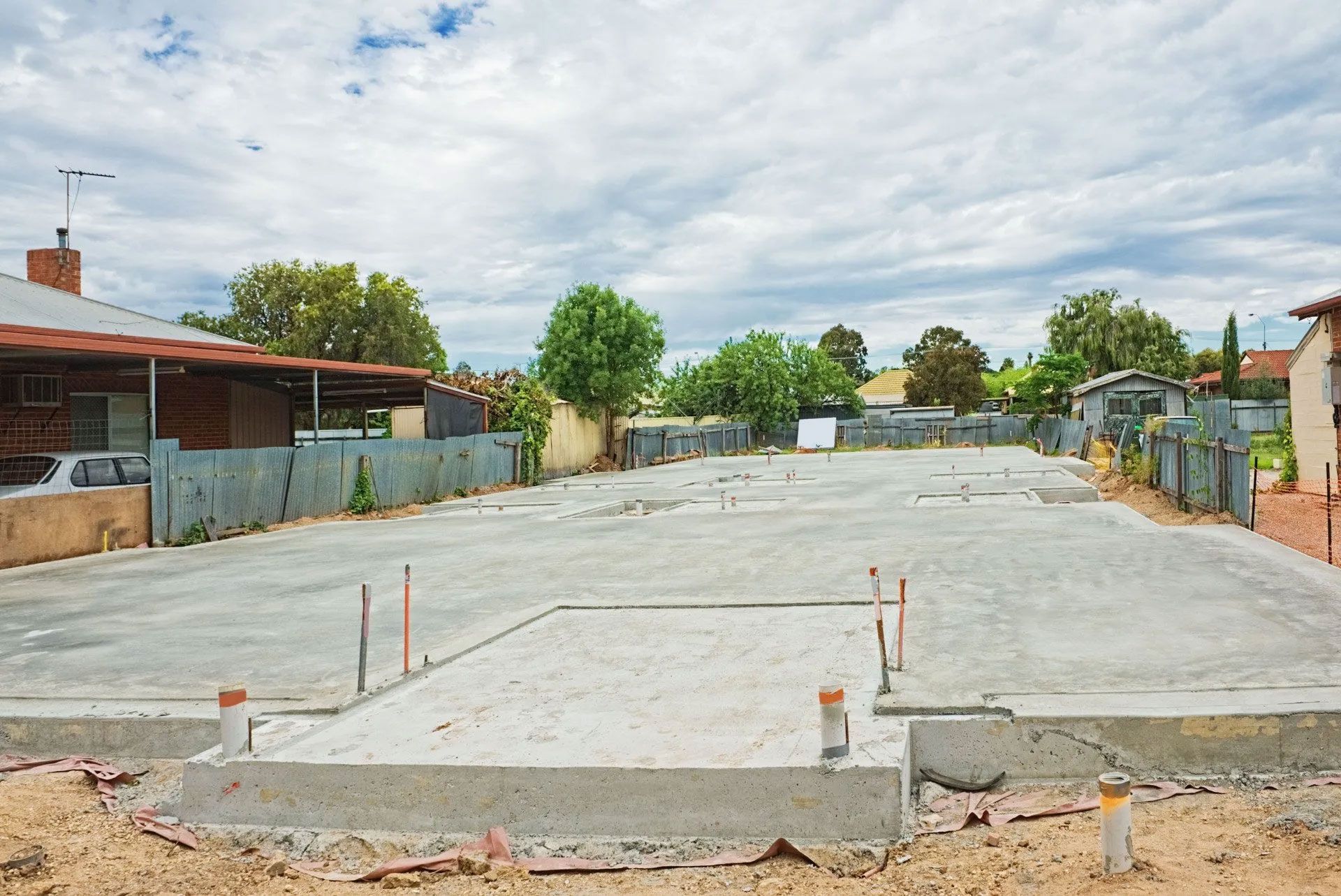 Concrete foundation for a building in a residential area, with metal fencing and cloudy sky.