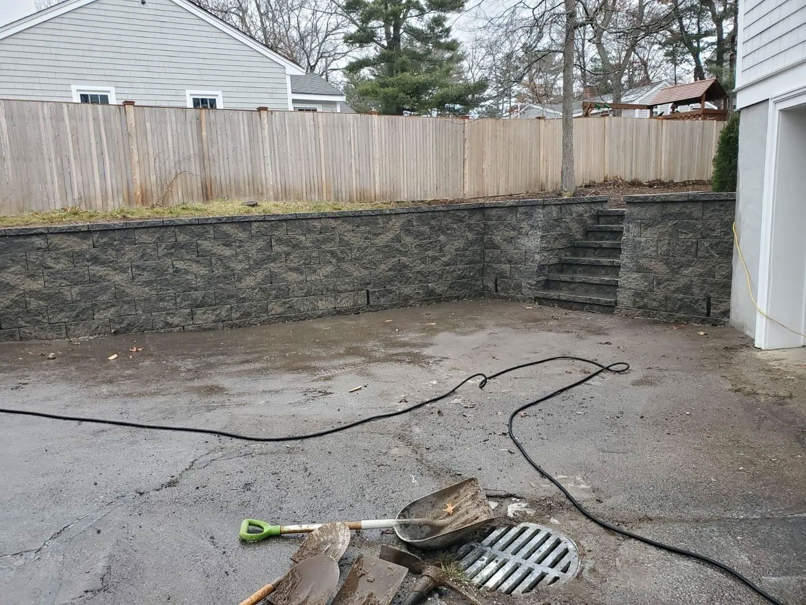 Black retaining wall, steps, and fence in a backyard with a drain and a shovel in foreground.