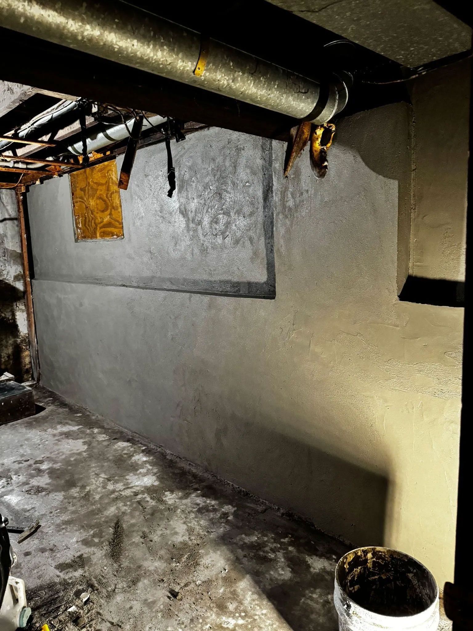 Basement wall with fresh concrete, pipes overhead, and a bucket on the floor.