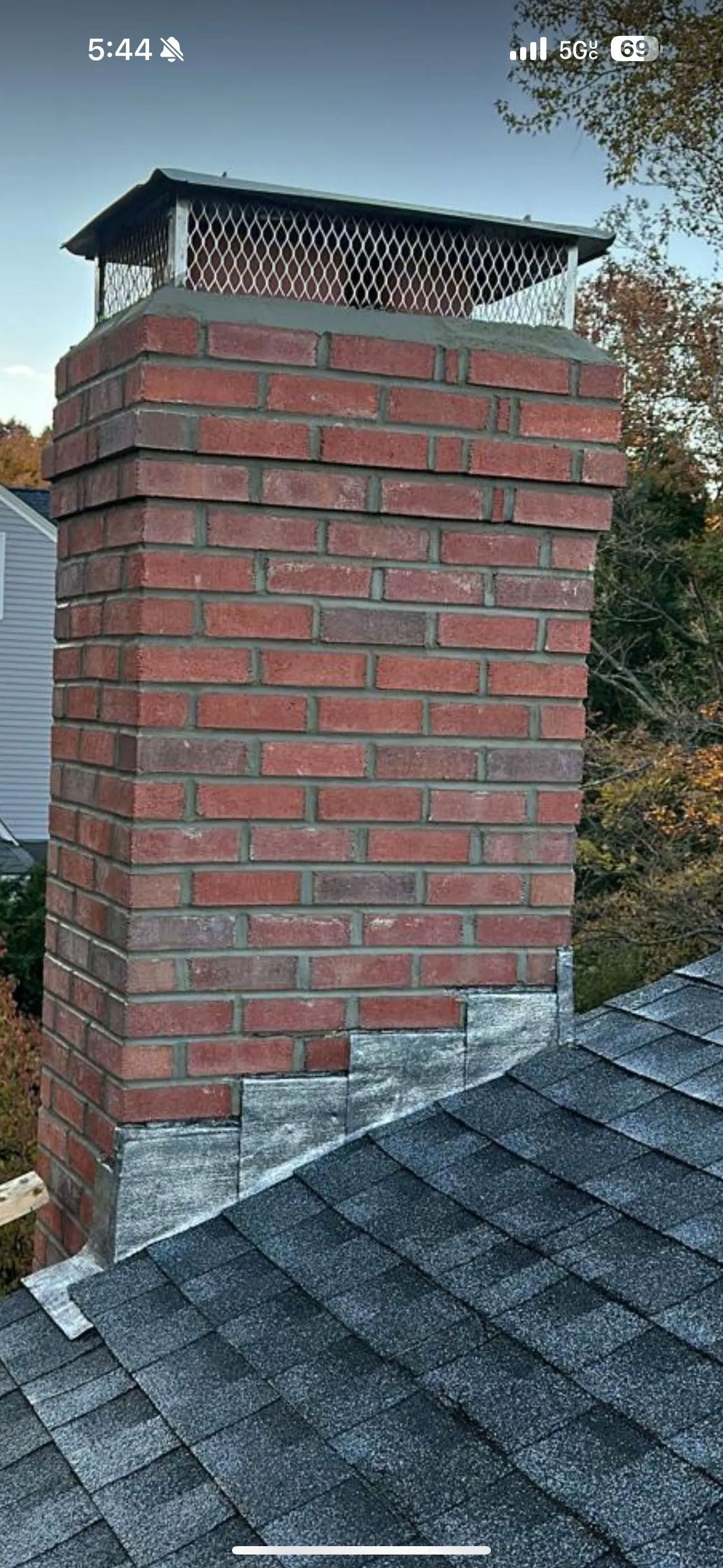 Brick chimney on a roof, with a metal mesh cap. Gray shingles, red bricks, blue sky.