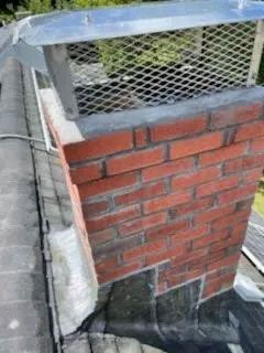 Brick chimney on a roof with a metal cap and mesh. Black sealant and shingles are also visible.