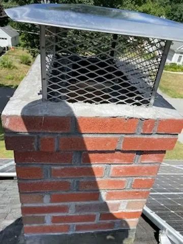 Red brick chimney with a metal mesh cap on a rooftop.