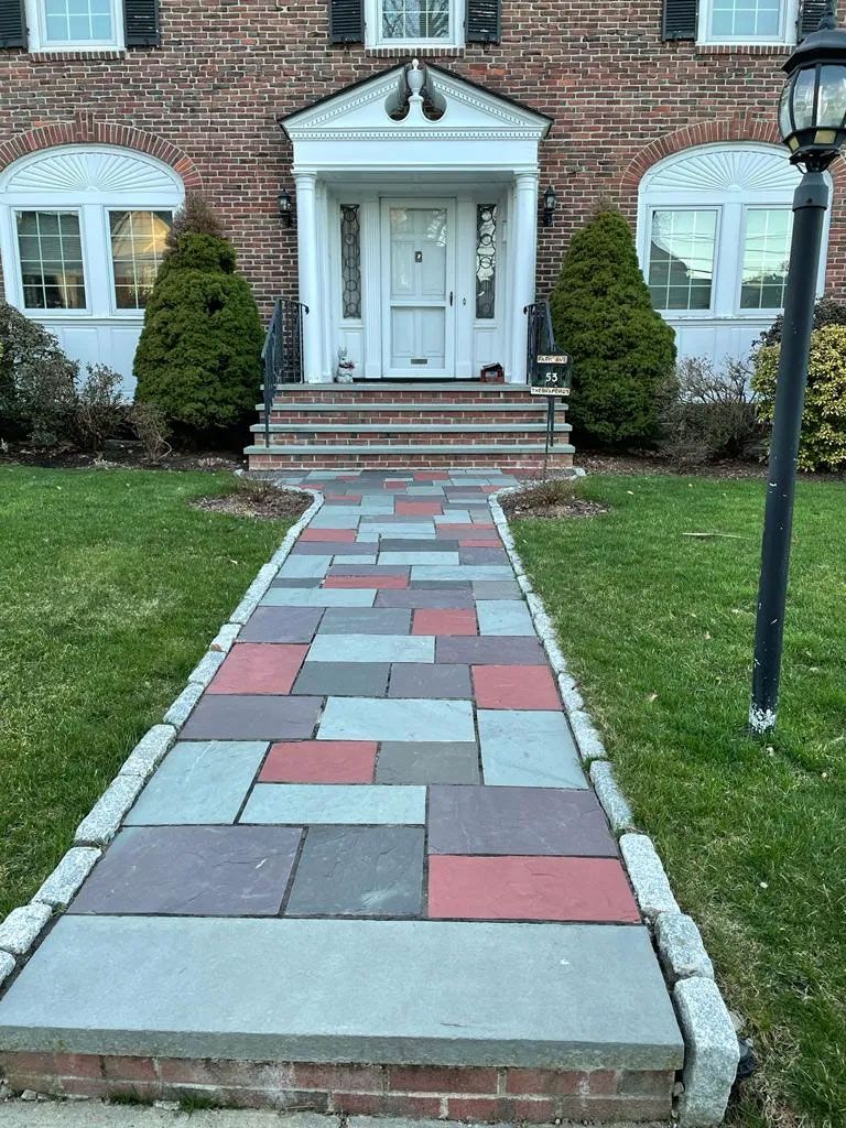 Brick house with a colorful paved walkway leading to the front door. Green lawn and bushes.