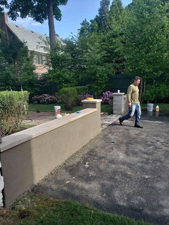 Man walks past a freshly plastered retaining wall in a backyard. Green trees and shrubs surround.