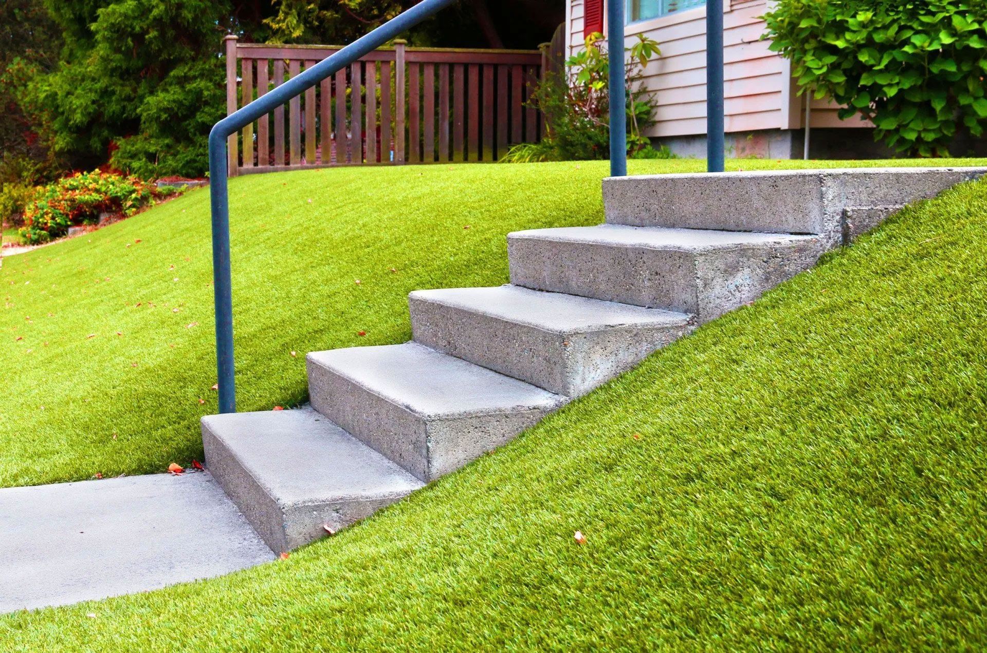 Concrete steps with a blue handrail on a grassy hill, leading to a house.