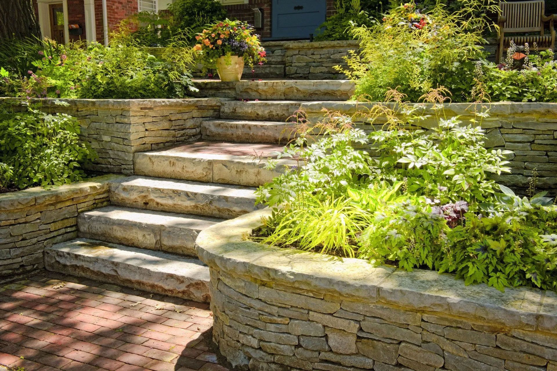 Stone steps and retaining walls in a garden, with brick path, lush plants, and a doorway in the background.