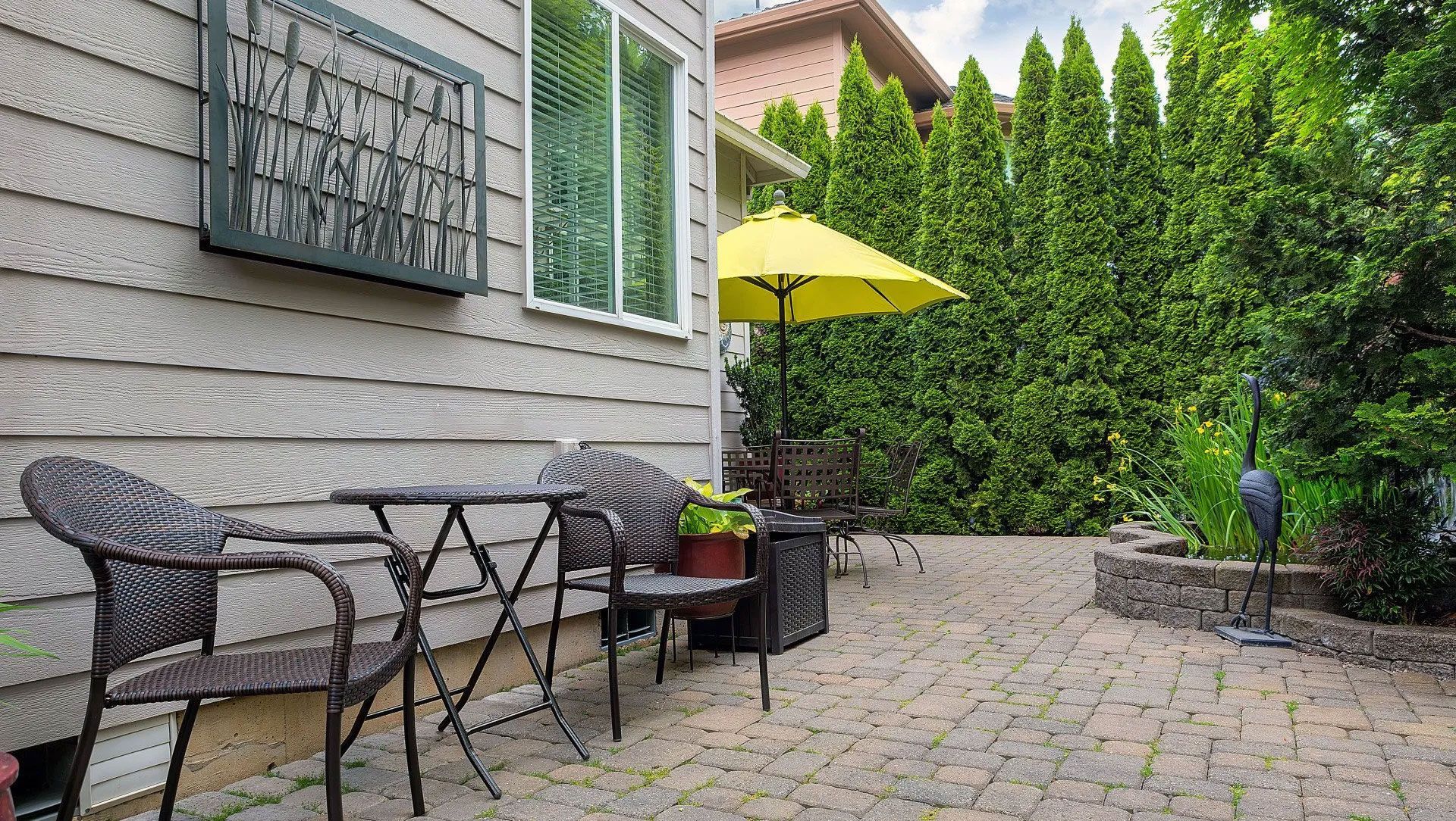 Patio with table, chairs, yellow umbrella, and hedge. House siding visible.